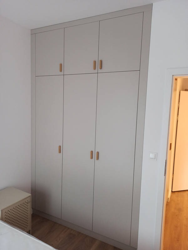 Jasnoszara szafa wnękowa z drewnianymi uchwytami, zabudowana od podłogi do sufitu, widoczna z boku w pokoju z drewnianą podłogą i fragmentem łóżka.