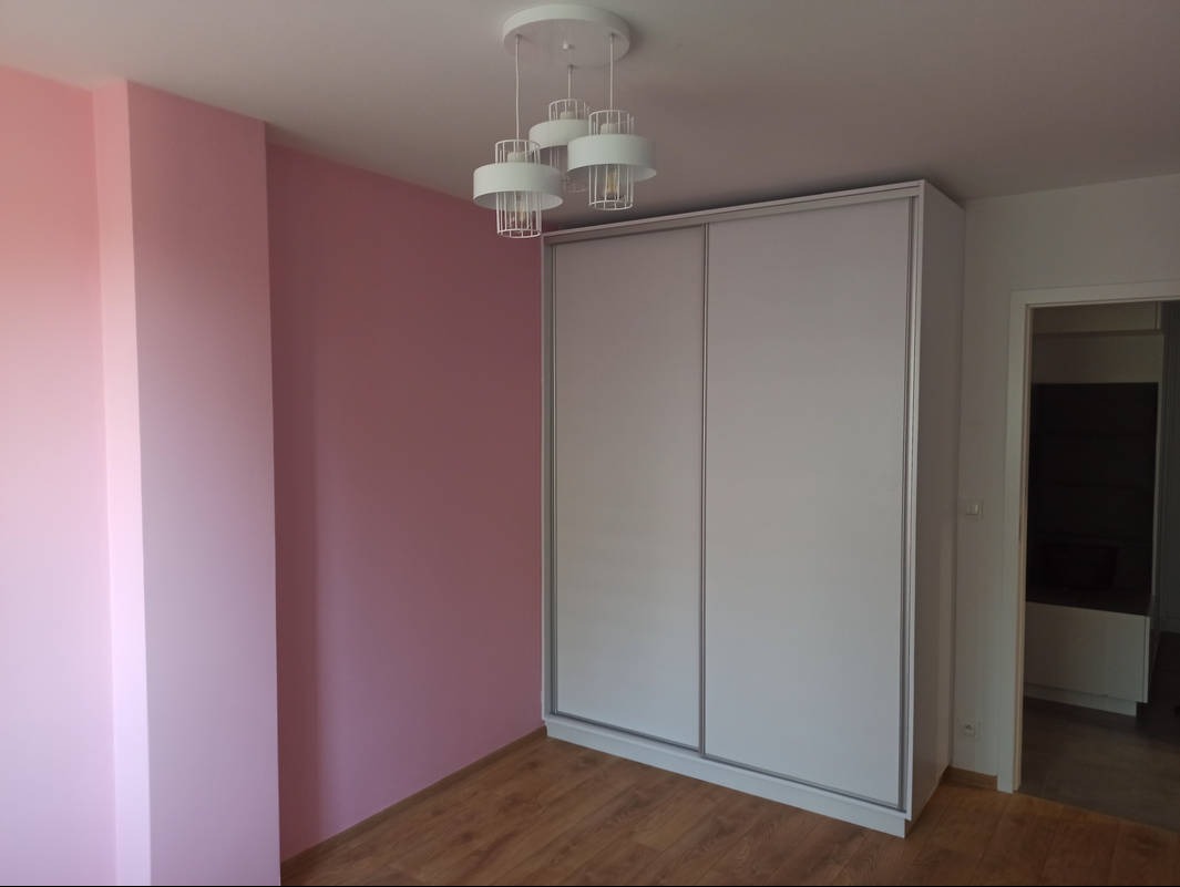 Jasna szafa przesuwna z trzema drzwiami w pokoju z różowymi ścianami i drewnianą podłogą. Widoczna nowoczesna lampa sufitowa.