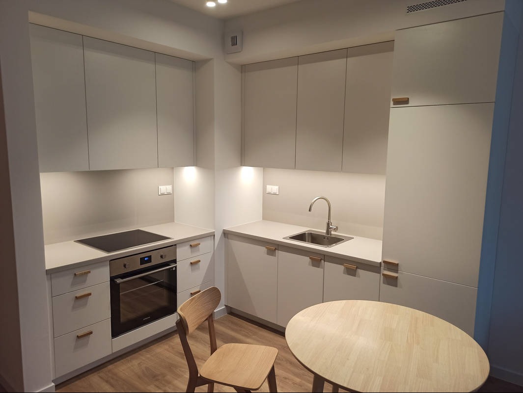 Jasna, nowoczesna kuchnia z drewnianymi akcentami: proste fronty, blat zintegrowany ze zlewem, płyta indukcyjna i drewniane krzesło przy okrągłym stole.