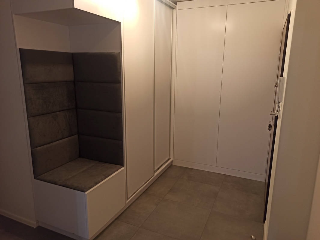 Biała szafa wnękowa z tapicerowanym siedziskiem i oświetleniem LED u góry. Minimalistyczny design, szara podłoga, drzwi przesuwne.