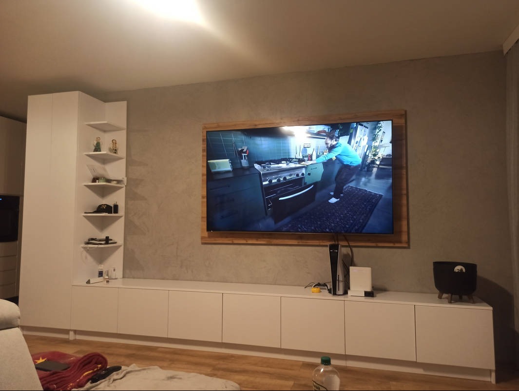 Biała, niska szafka RTV z blatem pod telewizorem w drewnianej ramie. Obok wysoka szafa z półkami narożnymi. Minimalistyczny design, szare ściany.