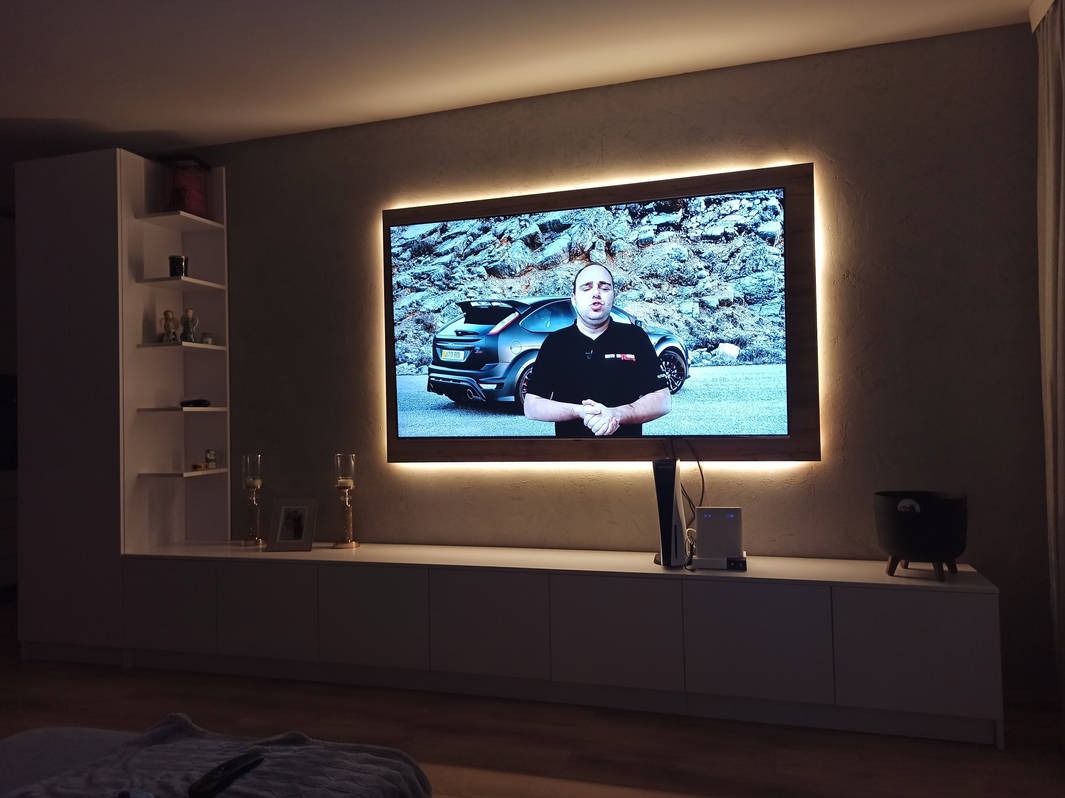 Biała szafka RTV z podświetlonym telewizorem, na którym widać mężczyznę i samochód. Obok regał z półkami i dekoracjami. Stonowana kolorystyka wnętrza.