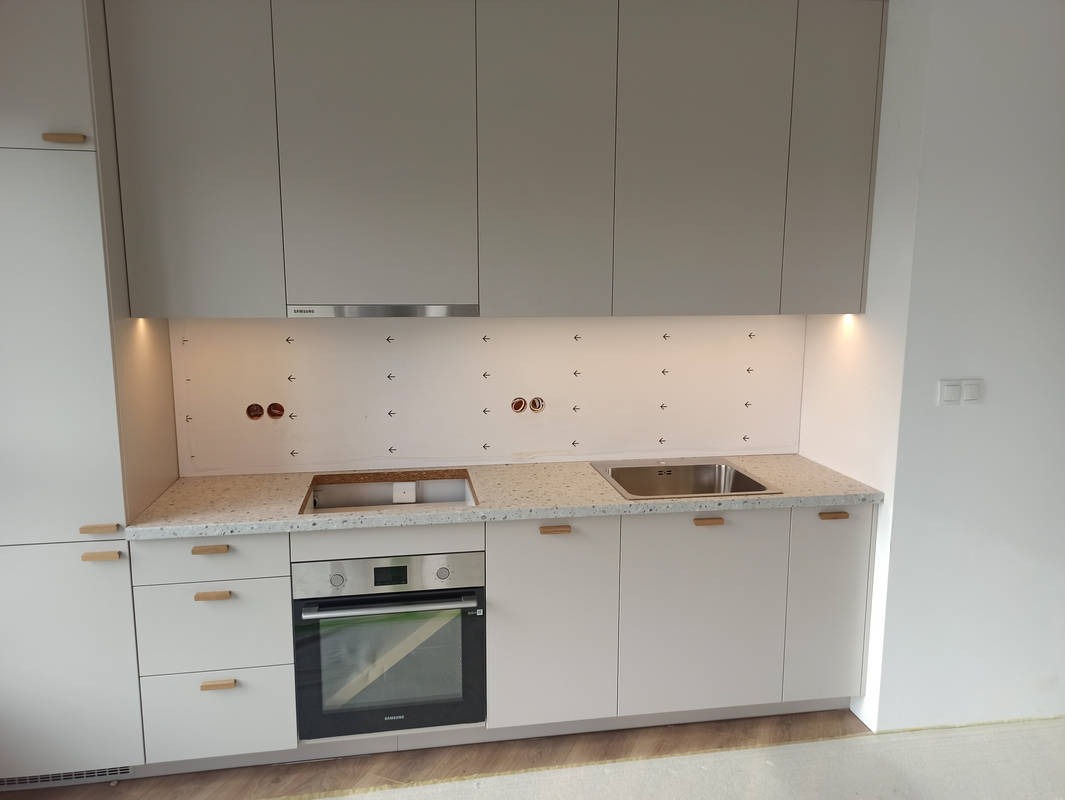 Jasna kuchnia z blatem w kropki, jeszcze bez wykończenia. Widoczne otwory na gniazdka i strzałki na ścianie nad blatem. Minimalistyczne uchwyty i piekarnik.