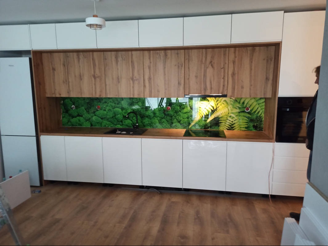 Nowoczesna kuchnia z białymi szafkami dolnymi i górnymi w kolorze drewna. Nad blatem panel z motywem roślinnym. Widoczny zlew i płyta grzewcza. Całość w jasnym pomieszczeniu.