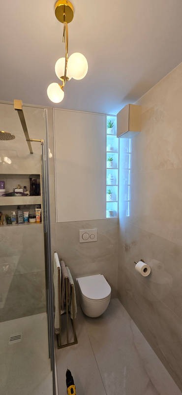 Nowoczesna łazienka z kabiną prysznicową, toaletą wiszącą i podświetlaną wnęką z roślinami. Złote akcenty oświetlenia i armatury. Na podłodze leży wkrętarka.