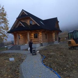 BARTOSZ KOSIOROWSKI - Nowo wybudowany drewniany dom z bali z czarnym dachem, widok z zewnątrz, z częściowo ułożoną ścieżką z kostki granitowej i minikoparką w tle, w mglisty dzień.