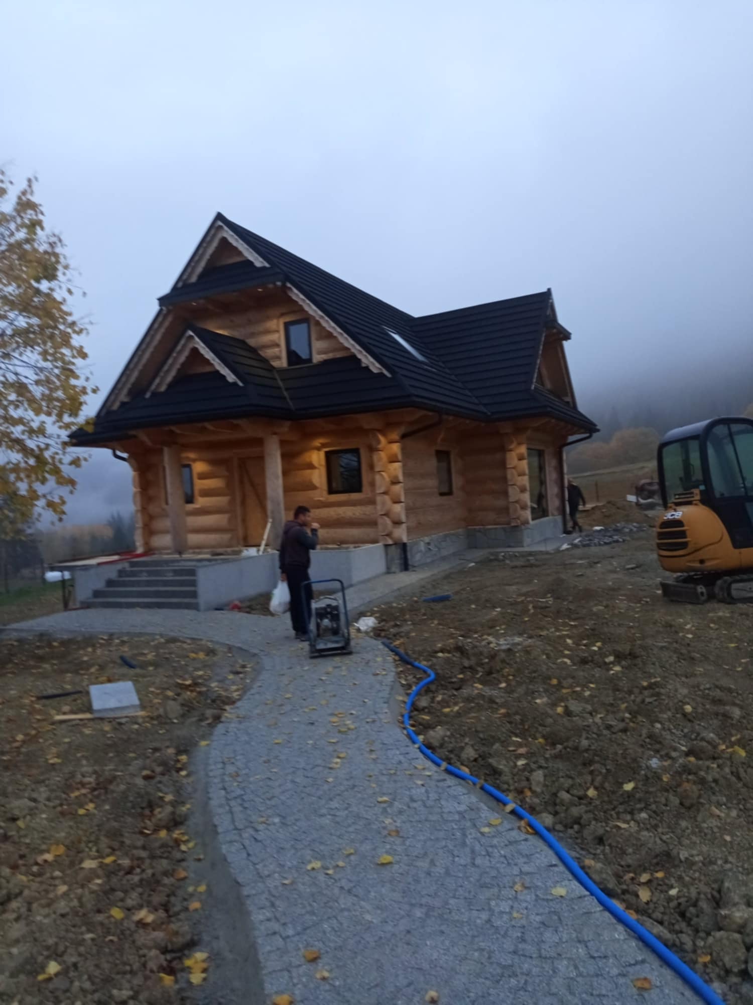 Nowo wybudowany drewniany dom z bali z czarnym dachem, widok z zewnątrz, z częściowo ułożoną ścieżką z kostki granitowej i minikoparką w tle, w mglisty dzień.
