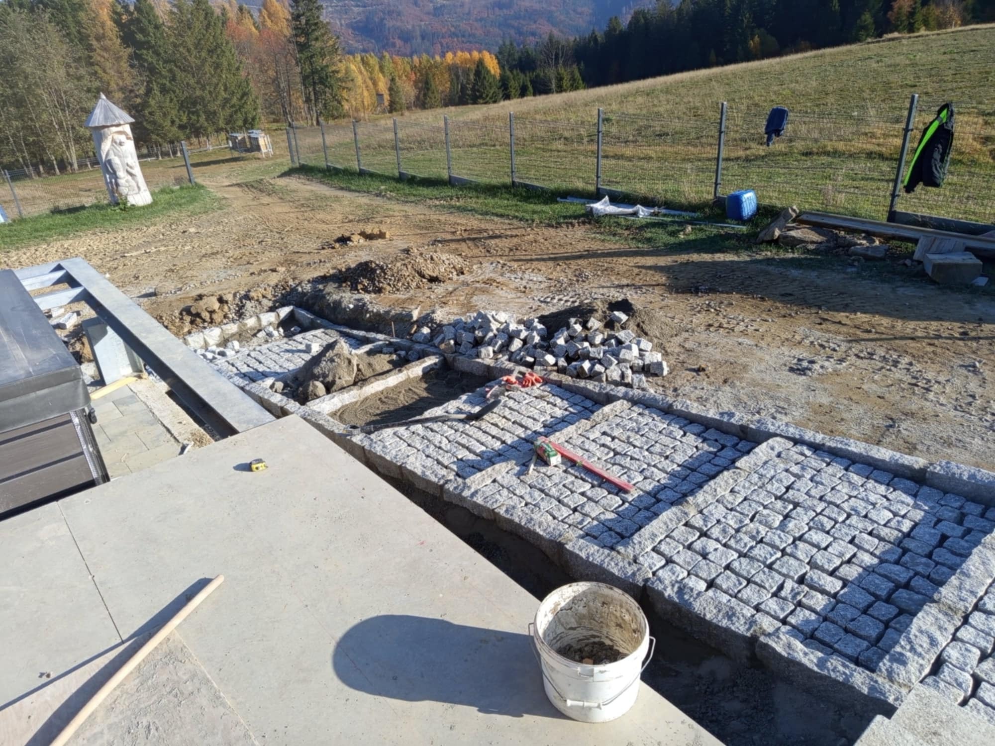 Fragment układanej nawierzchni z kostki granitowej na tle krajobrazu górskiego z widoczną rzeźbą plenerową, narzędziami i zapasem kostki.