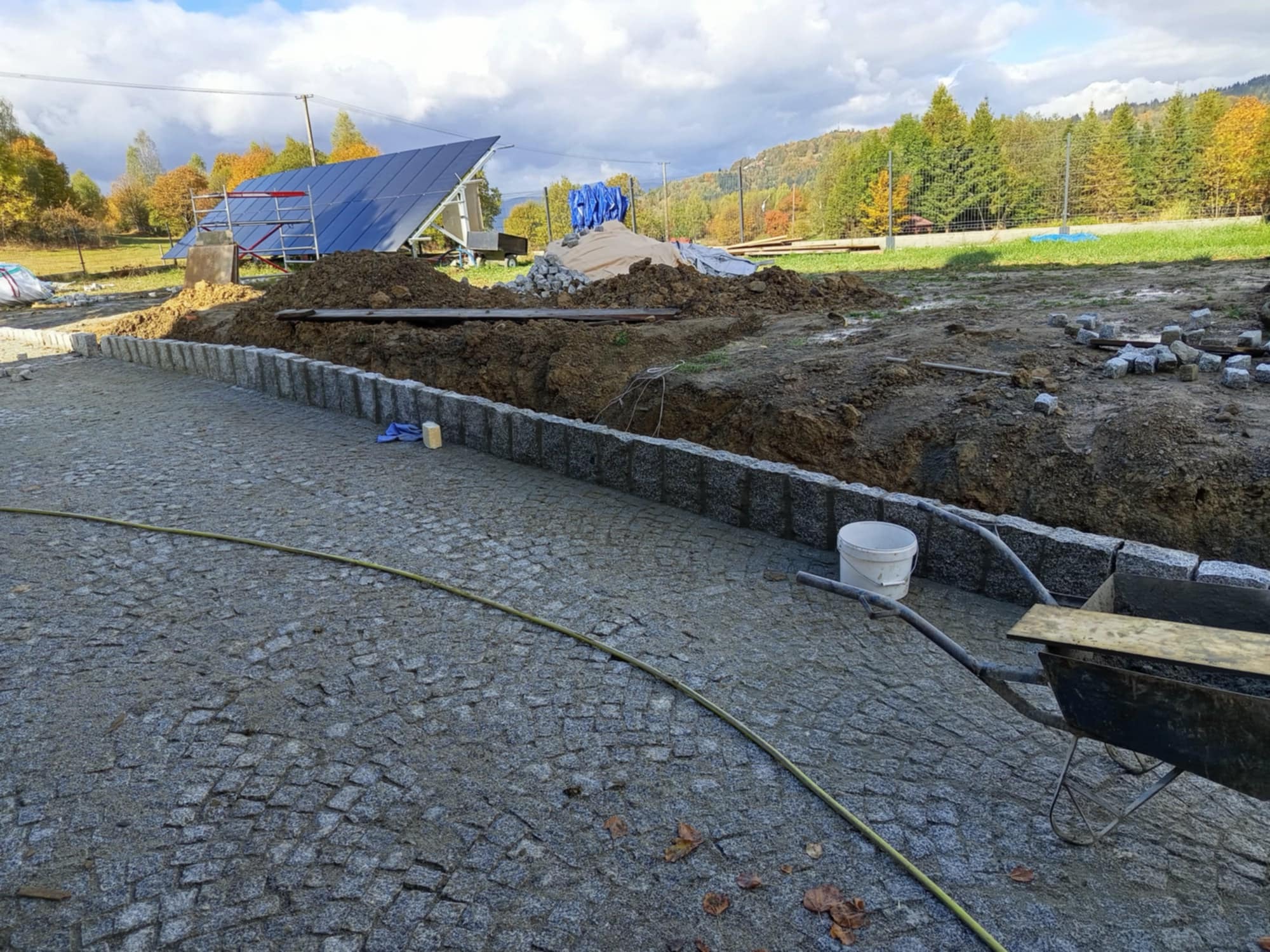 Świeżo ułożona nawierzchnia z kostki granitowej z obrzeżami, widoczny fragment wykopu, taczka, wiadro i panele słoneczne w tle na tle pagórkowatego krajobrazu.