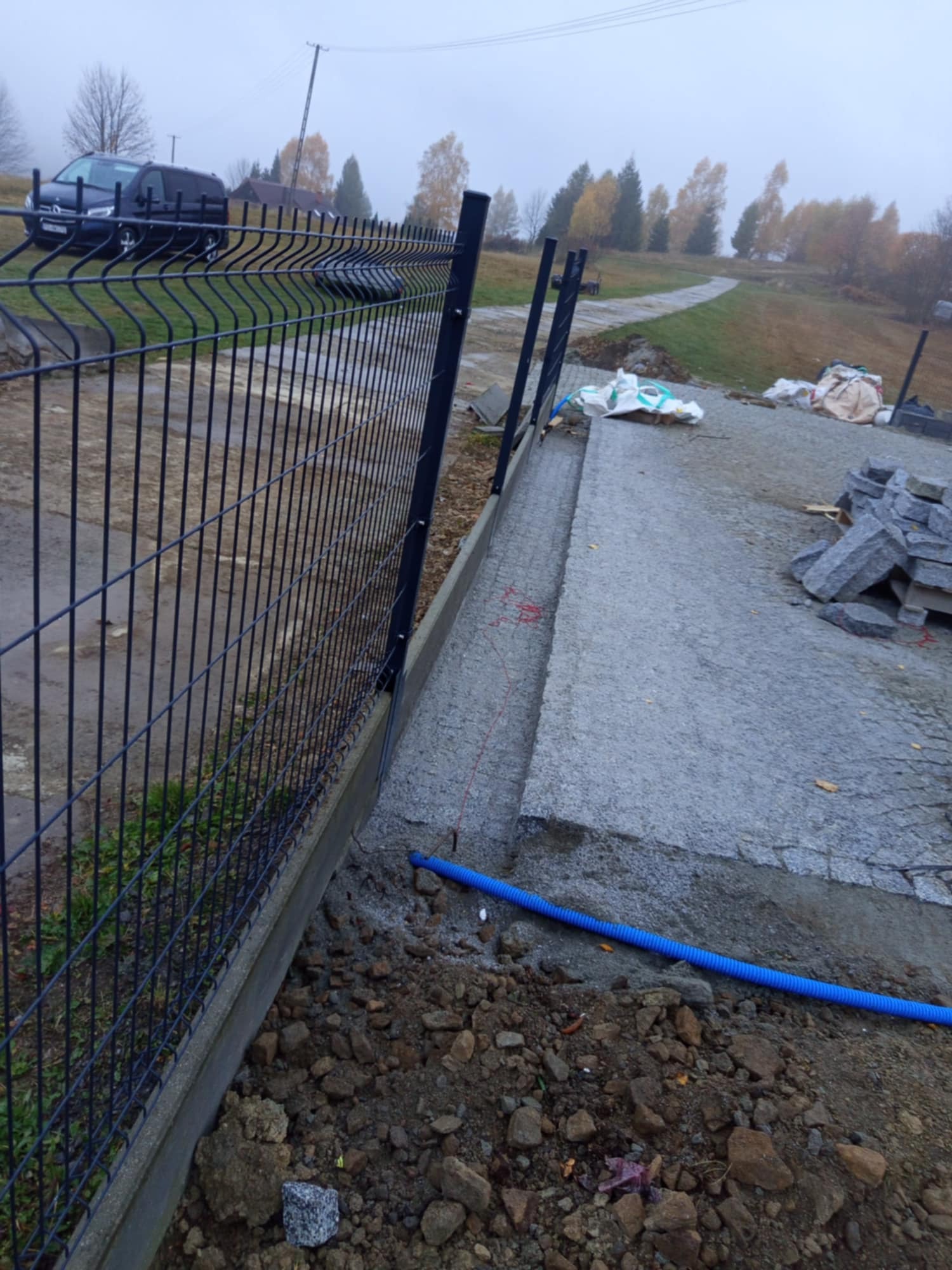 Montaż ogrodzenia panelowego i układanie kamiennej nawierzchni na podbudowie betonowej, widoczne worki z materiałami i niebieski przewód instalacyjny.