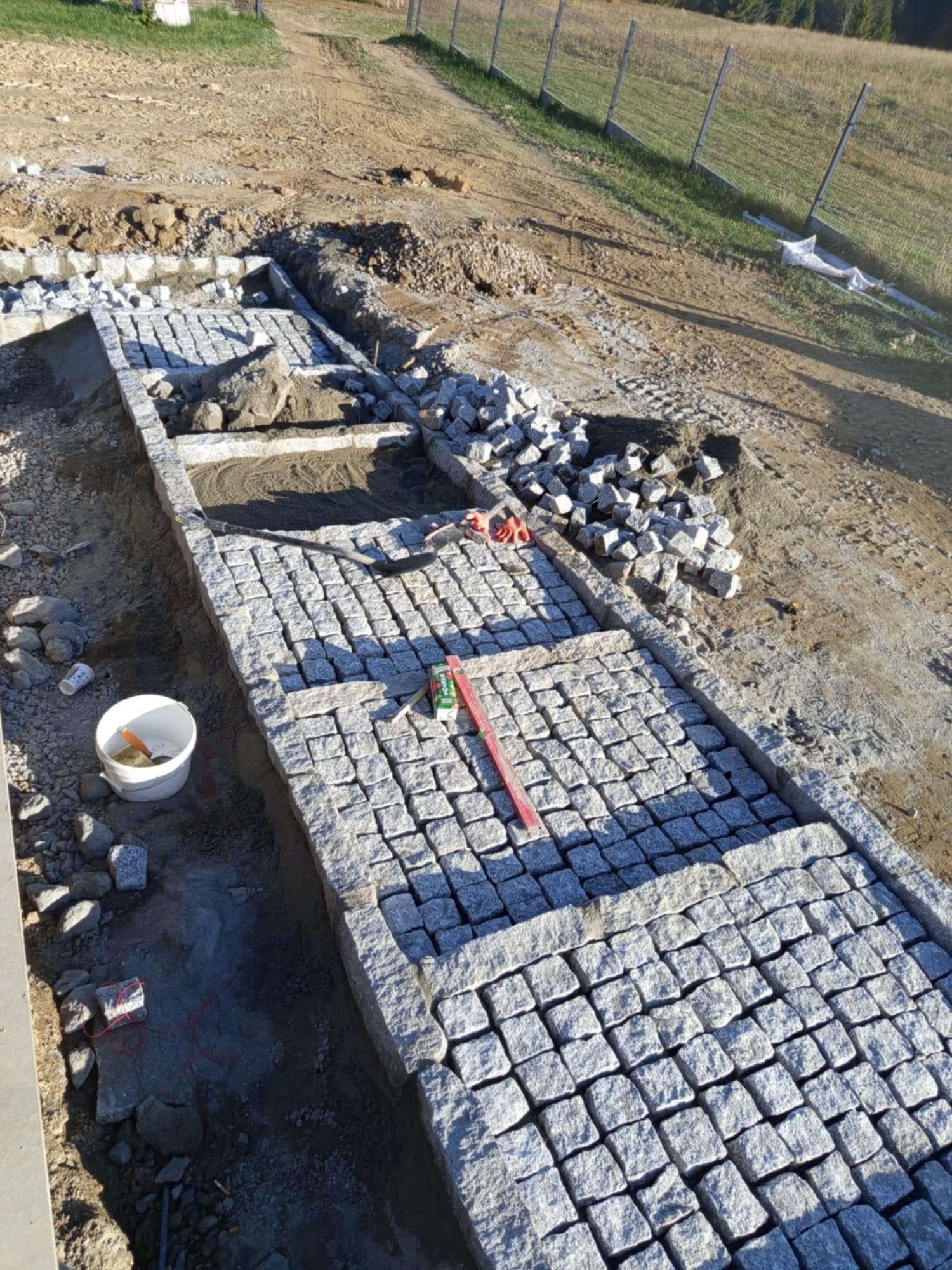 Układanie granitowej kostki brukowej w trakcie budowy ścieżki, widoczny poziomica, kubeł z zaprawą i zapas kostki, tło z ziemią i siatką ogrodzeniową.