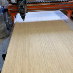 Obróbka CNC płyty bambusowej, frezarka z pomarańczową ramą. Widoczne detale frezowania i tekst na płycie. Obróbka na potrzeby mebli na wymiar.