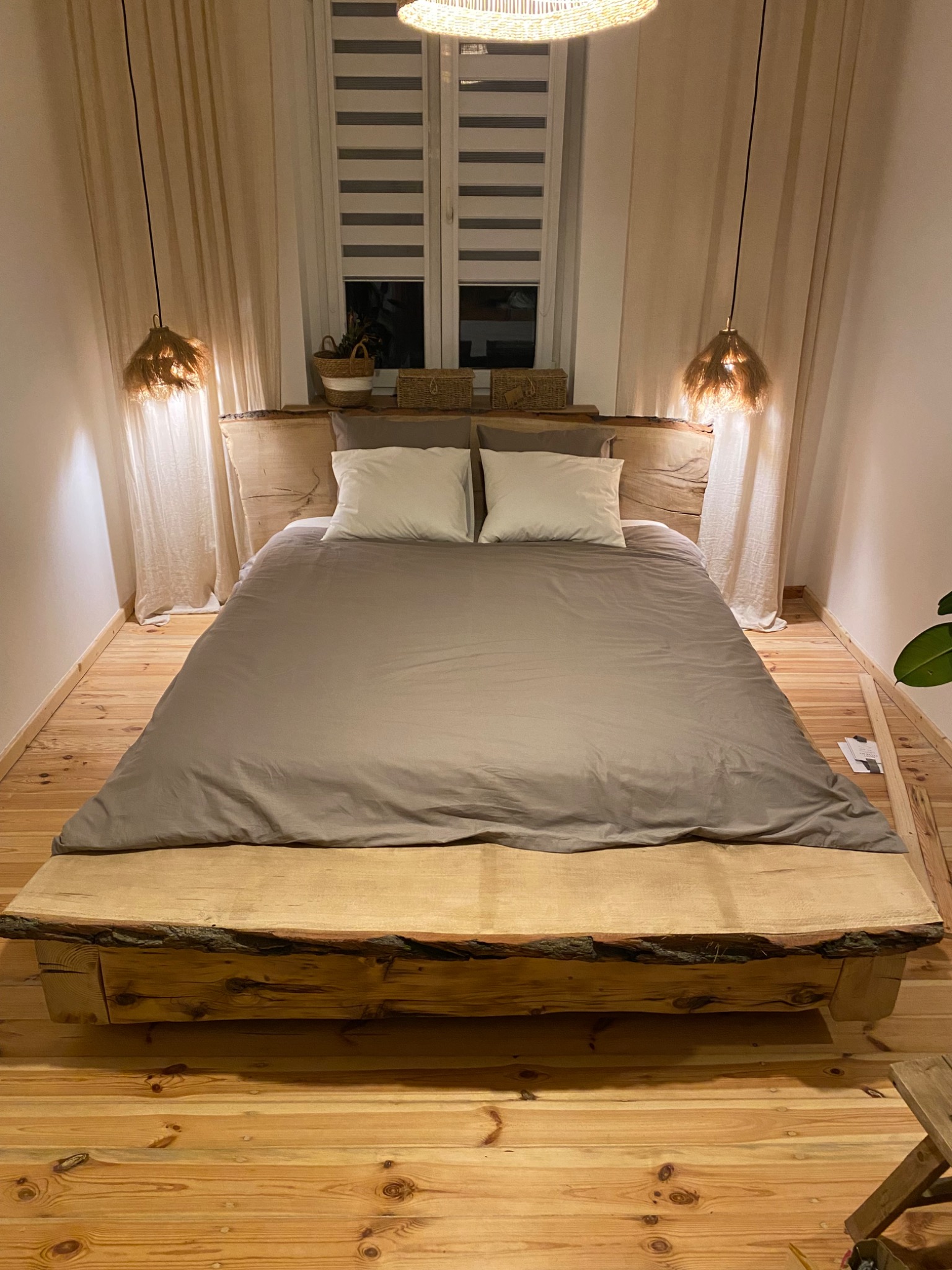 Drewniane łóżko z naturalnymi krawędziami, podwieszane lampy z abażurami z rafii, drewniana podłoga i jasne zasłony tworzą przytulną sypialnię w stylu rustykalnym.