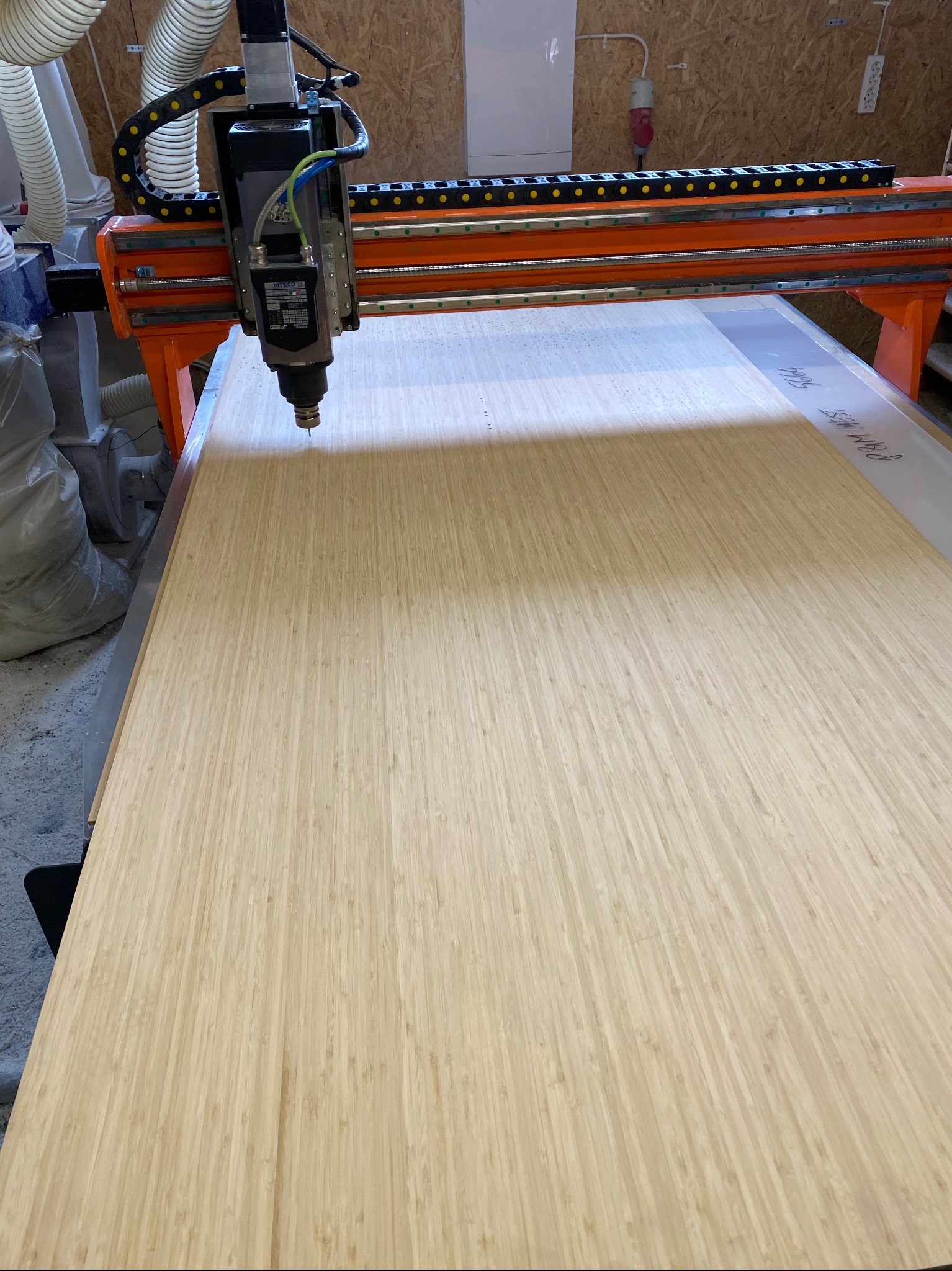Obróbka CNC płyty bambusowej, frezarka z pomarańczową ramą. Widoczne detale frezowania i tekst na płycie. Obróbka na potrzeby mebli na wymiar.