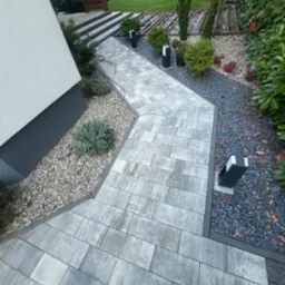 Ścieżka z szarych prostokątnych płyt betonowych, obrzeża z ciemnego kamienia, oświetlenie ogrodowe i dekoracyjny żwir wzdłuż ścieżki.