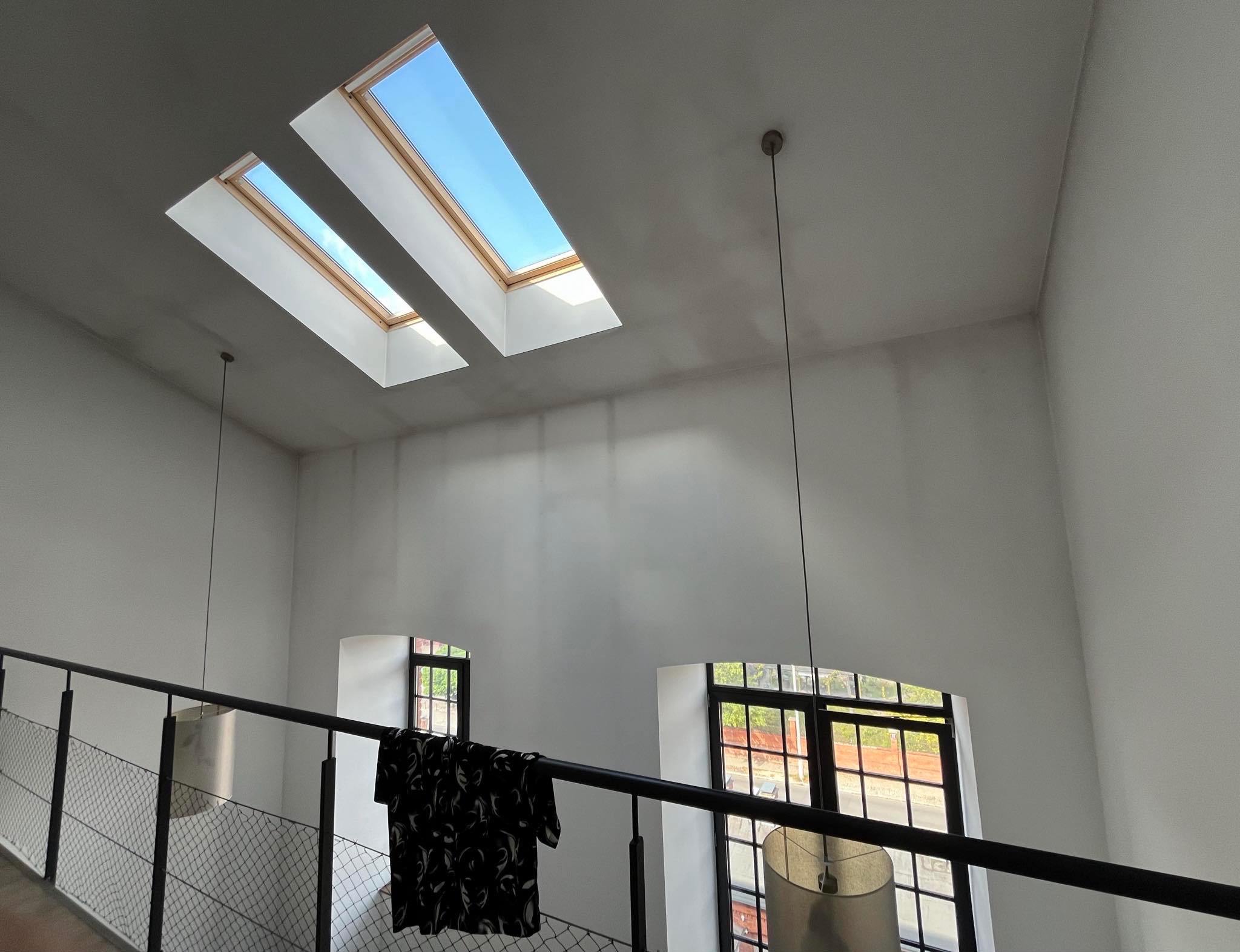 Wnętrze po odświeżeniu: białe ściany, dwa okna dachowe z widokiem na niebo, lampy wiszące, czarna balustrada schodów z siatką, na balustradzie powieszona czarno-biała koszula.