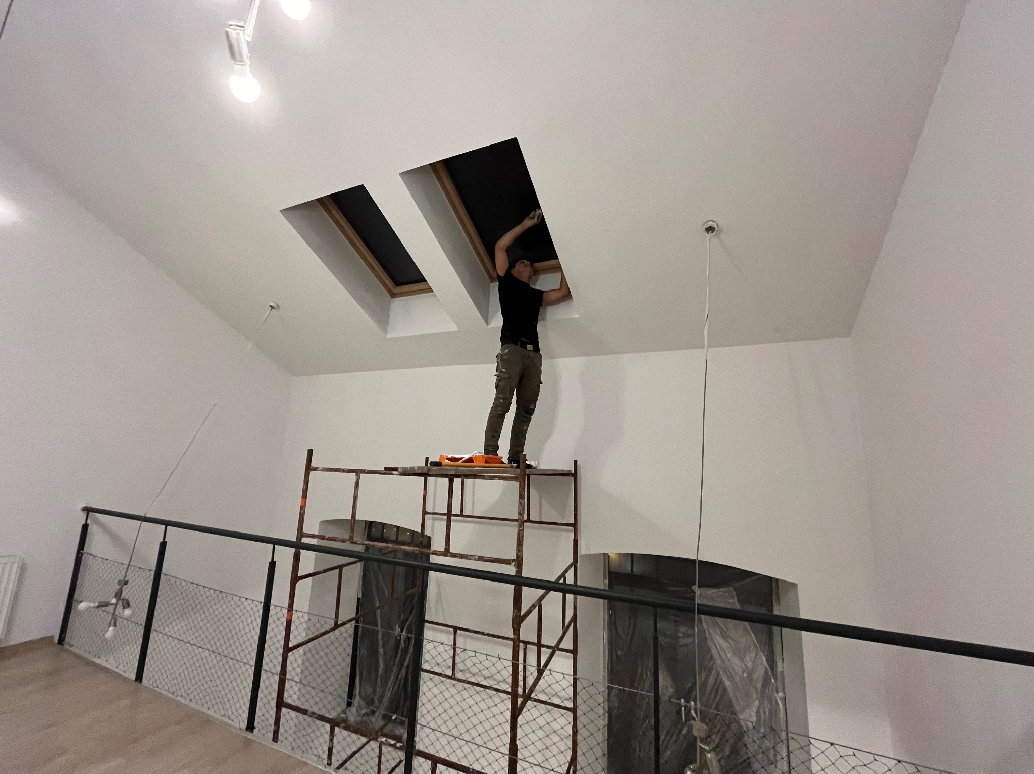 Pracownik na rusztowaniu maluje ramę okna dachowego w nowym, wysokim pomieszczeniu, widoczne dwa okna i białe ściany.