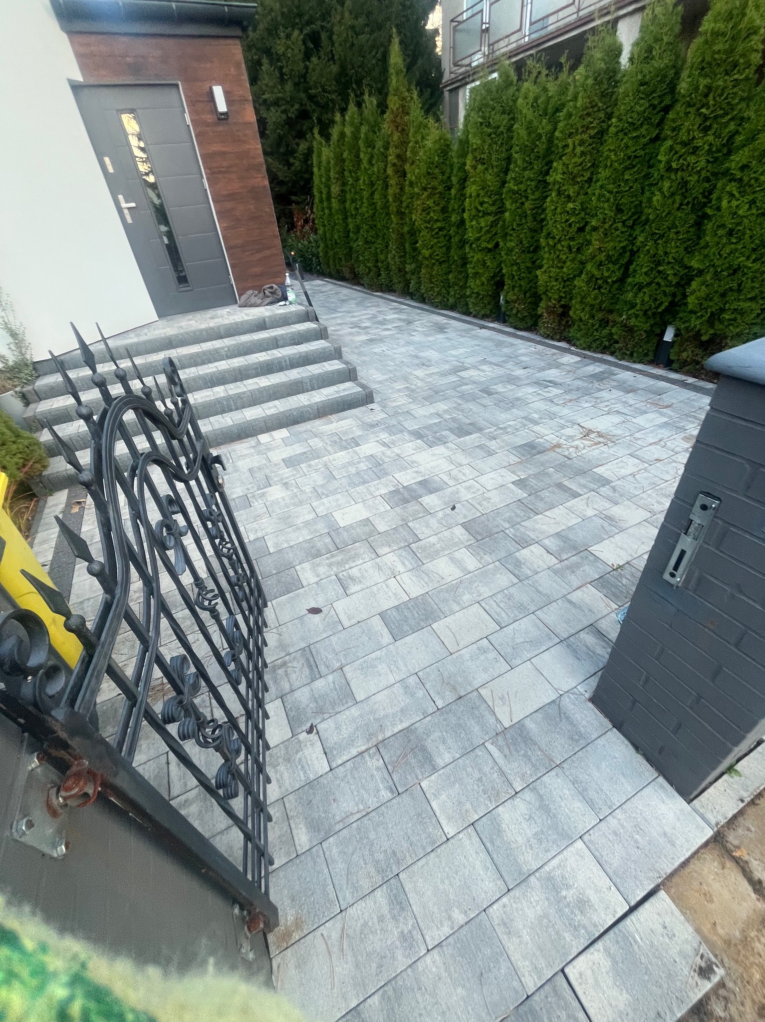 Ułożona kostka granitowa w odcieniach szarości prowadząca od bramy z ozdobnymi elementami do schodów wejściowych domu z ciemnymi ramami okiennymi i drzwiami, obok rząd gęstych, wysokich tui.