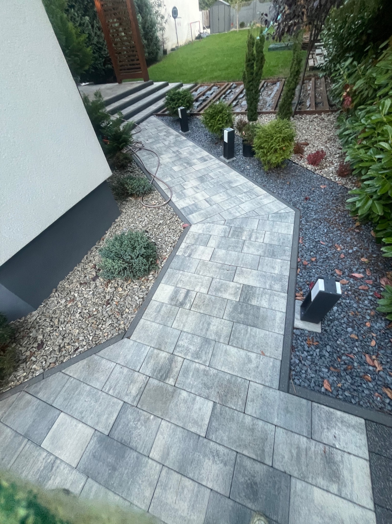 Ścieżka z szarych prostokątnych płyt betonowych, obrzeża z ciemnego kamienia, oświetlenie ogrodowe i dekoracyjny żwir wzdłuż ścieżki.