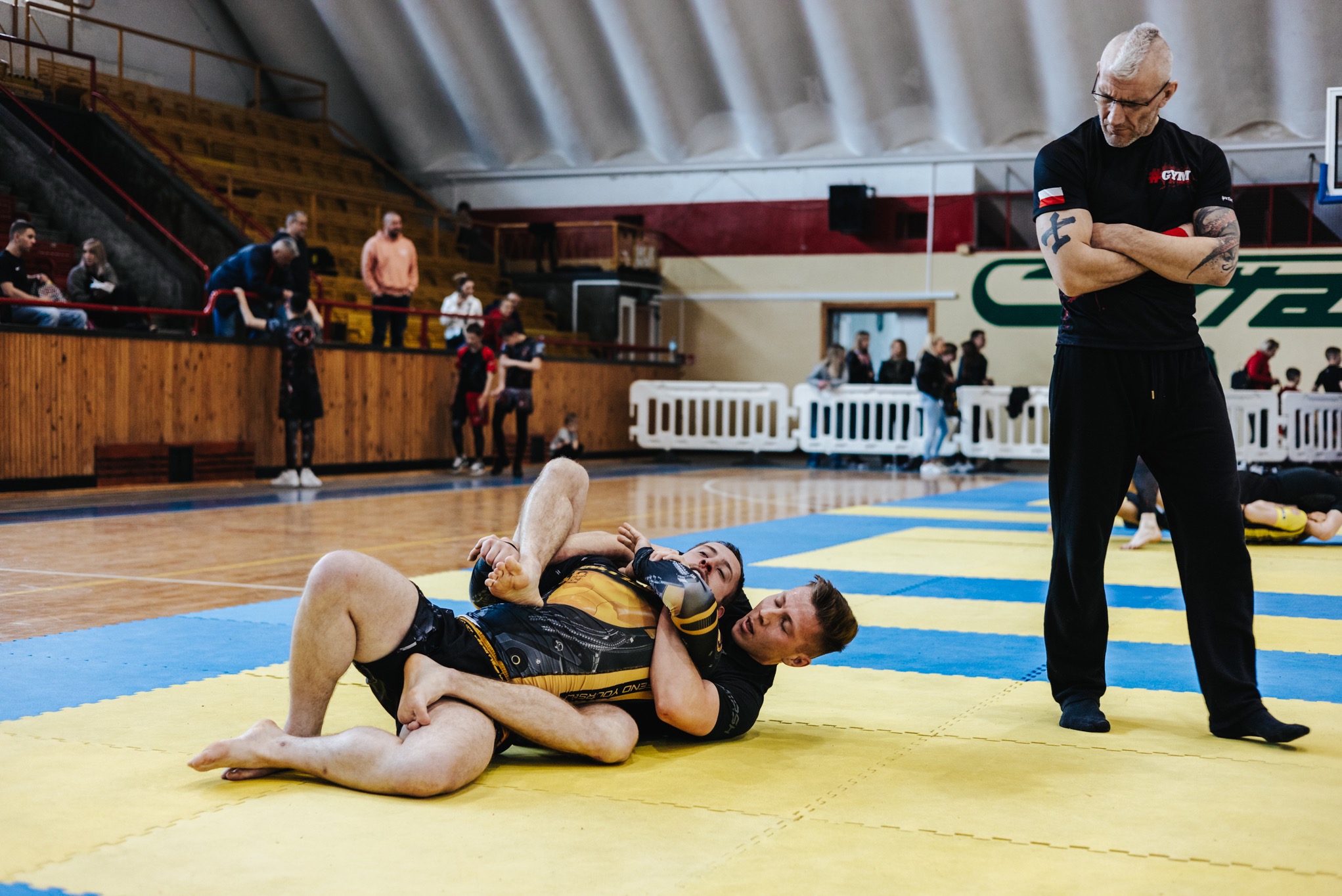 Trener personalny nadzoruje sparing MMA na macie w hali sportowej. Zawodnicy w kimonach, z widownią w tle i sędzią z tatuażami na ramionach.