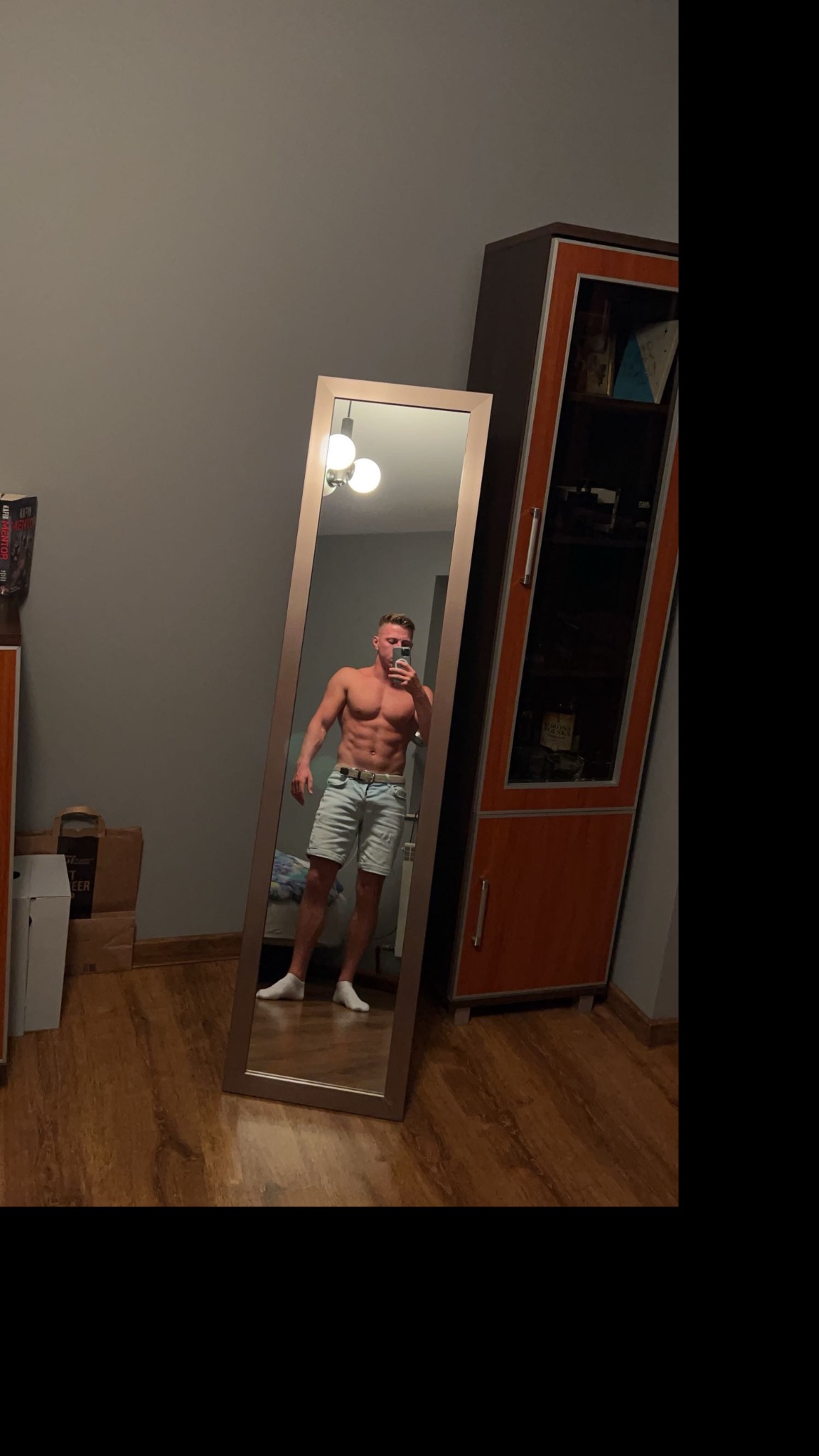 Mężczyzna z wyraźnie zarysowanymi mięśniami brzucha, pozujący do zdjęcia w lustrze w pomieszczeniu z drewnianą podłogą i szafką w tle. Widoczna lampa sufitowa.