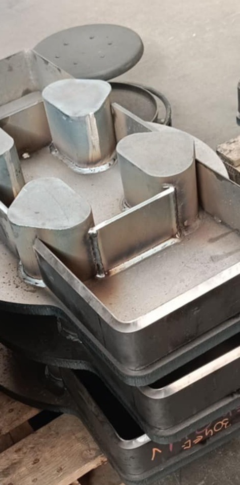 Metalowa konstrukcja z elementami spawania, widoczne spoiny i połączenia, surowy metal, częściowo pomalowana na czarno, ułożona na drewnianej palecie.