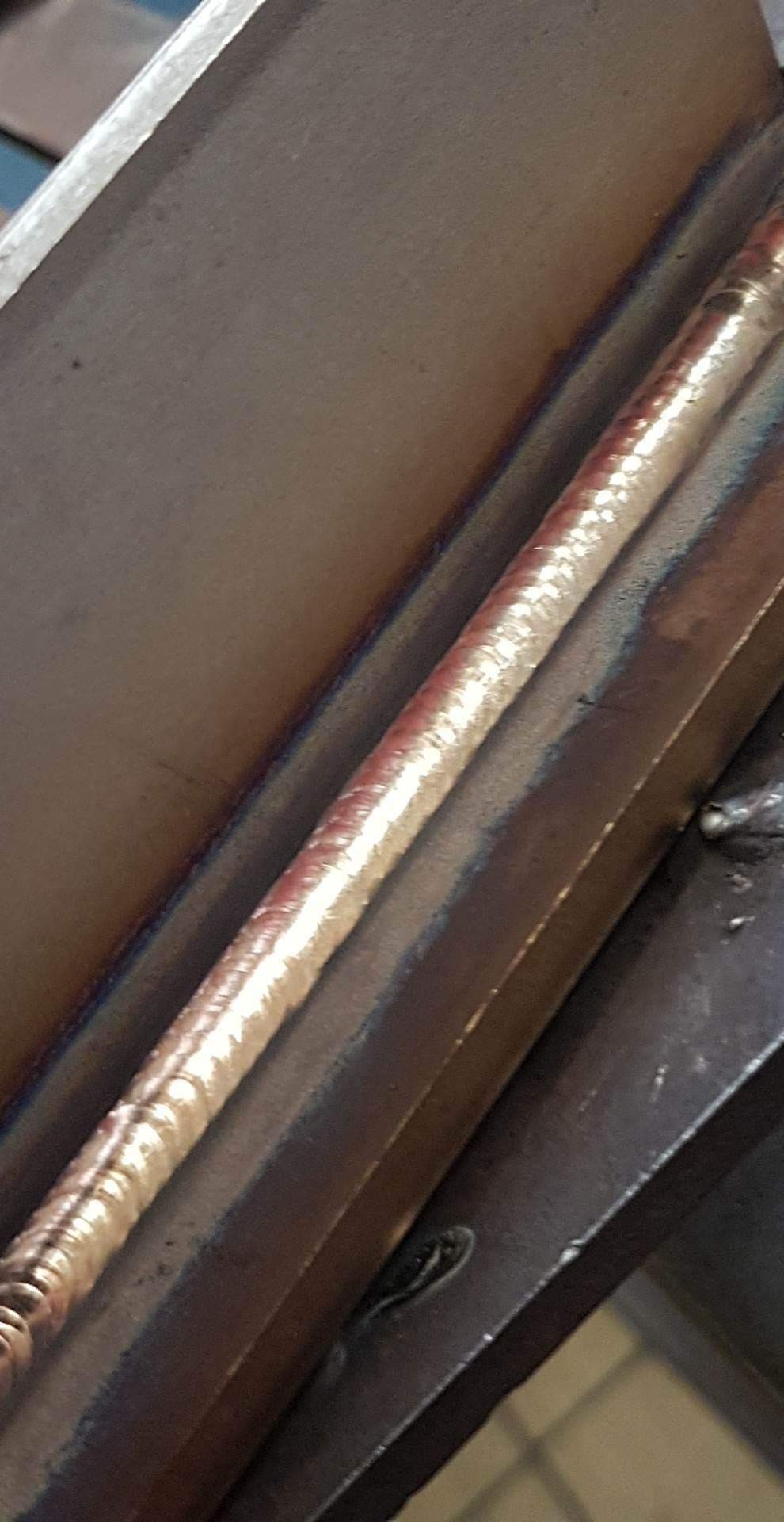 Połączenie spawane na metalowej konstrukcji, widoczny szew o nieregularnej fakturze i opalizujących kolorach, z bliska.