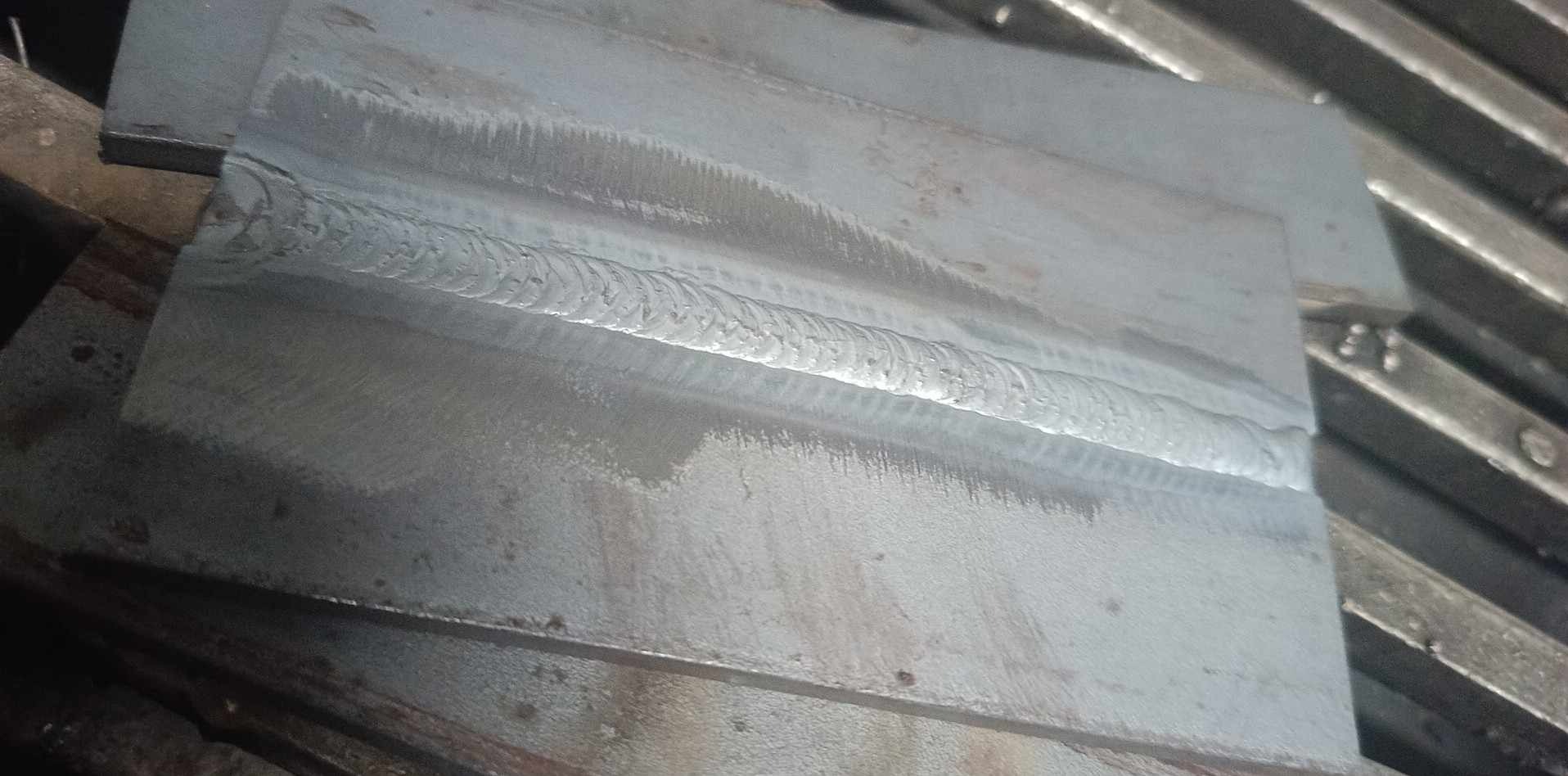 Spoina na dwóch złączonych, szarych, metalowych płytach, widoczne nierówności i przebarwienia na powierzchni metalu.