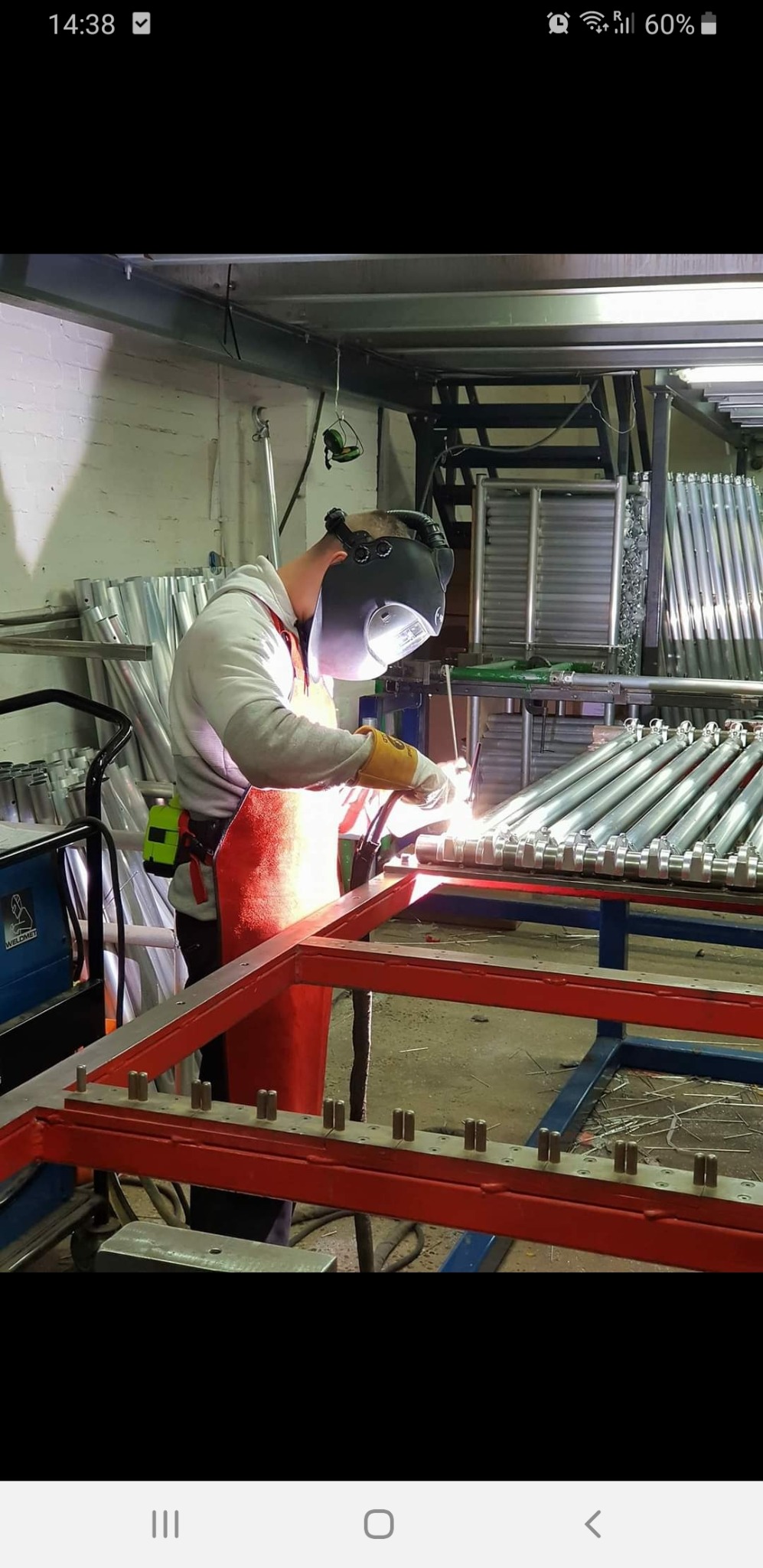 Pracownik w masce spawalniczej i fartuchu spawa metalową konstrukcję w warsztacie, w tle widoczne rusztowania.