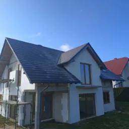 Dom jednorodzinny w trakcie budowy z częściowo ukończonym, ciemnoszarym dachem z dachówki ceramicznej, widoczne rusztowania i ocieplenie styropianowe na ścianach, błękitne niebo w tle.