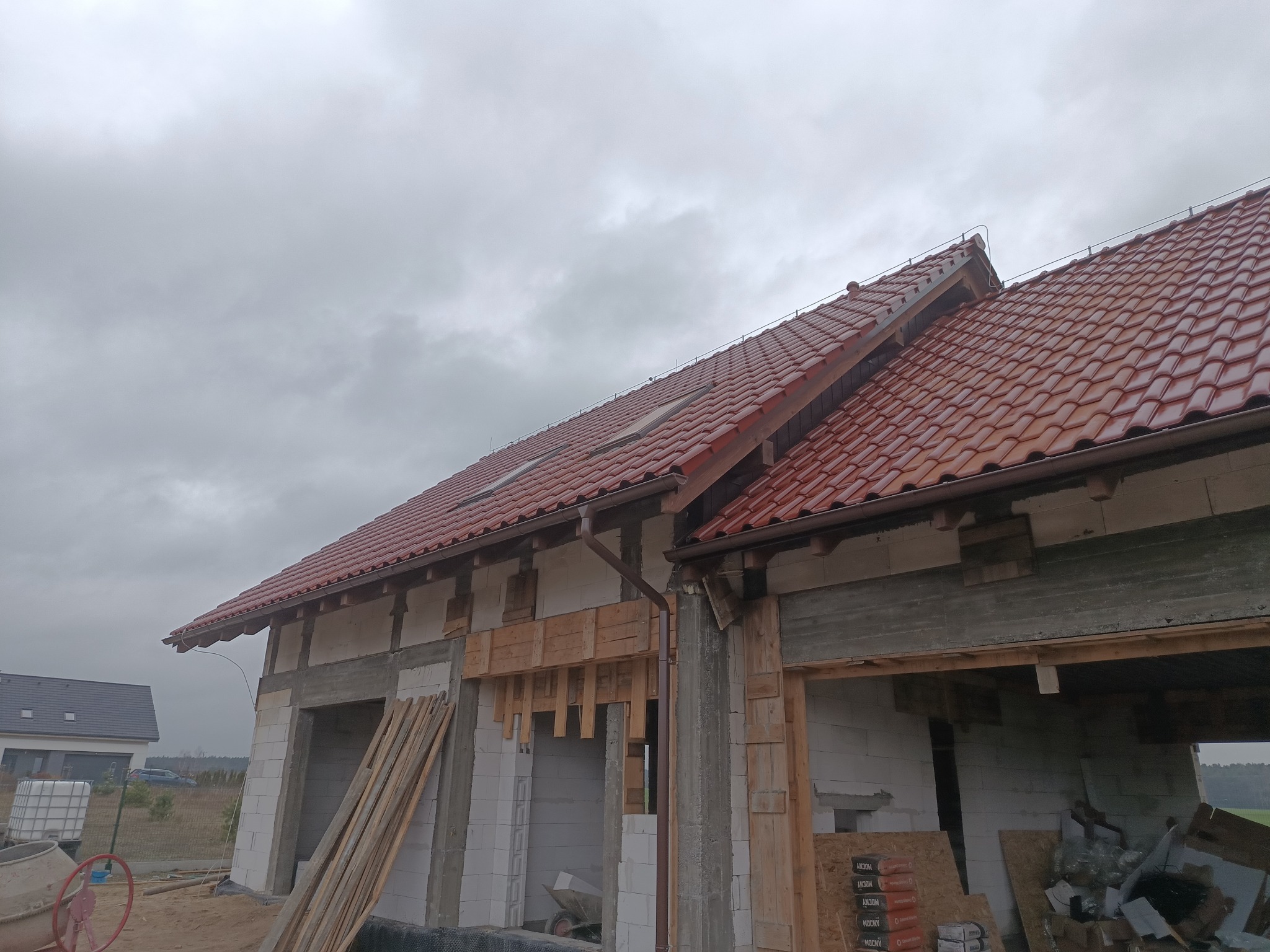 Nowy, błyszczący dach z czerwonej dachówki ceramicznej na budynku w trakcie budowy, widoczne rynny i elementy konstrukcyjne dachu, w tle pochmurne niebo.