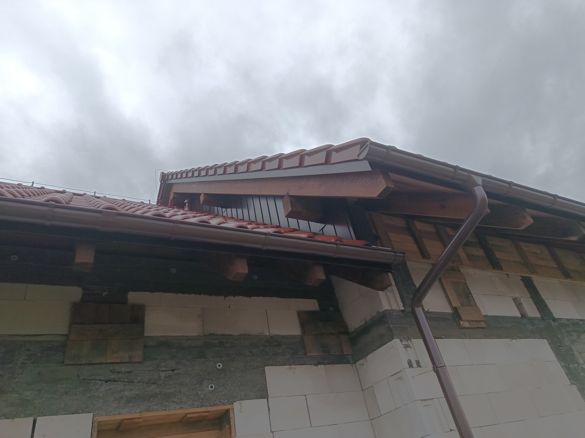 Fragment nowo budowanego domu z widocznym dachem pokrytym czerwoną dachówką, brązowymi rynnami i surową ścianą z betonu komórkowego na tle pochmurnego nieba.