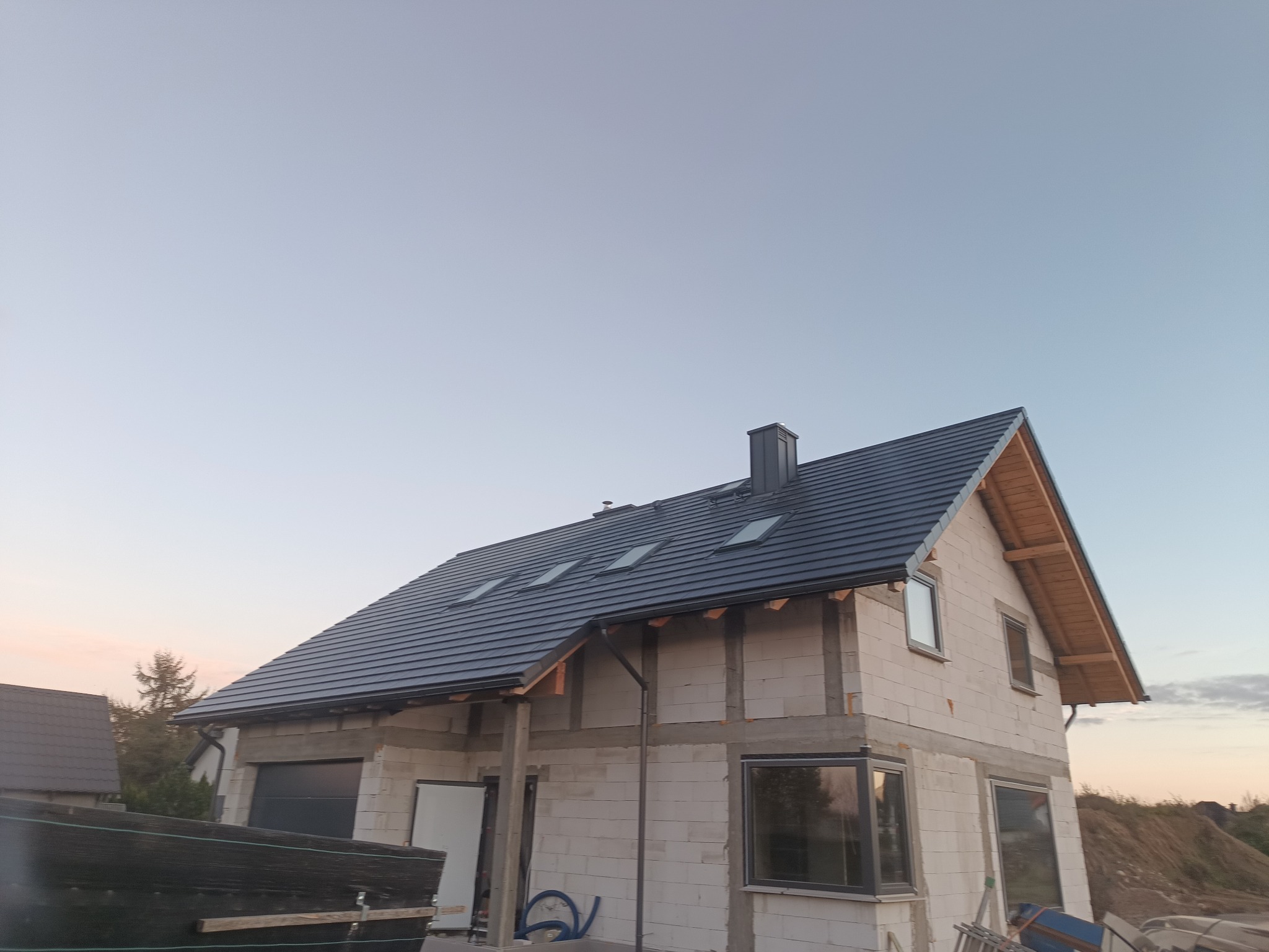 Dom w trakcie budowy z nowym, ciemnoszarym dachem z dachówką ceramiczną i oknami dachowymi, widoczne orynnowanie, ściany z betonu komórkowego.