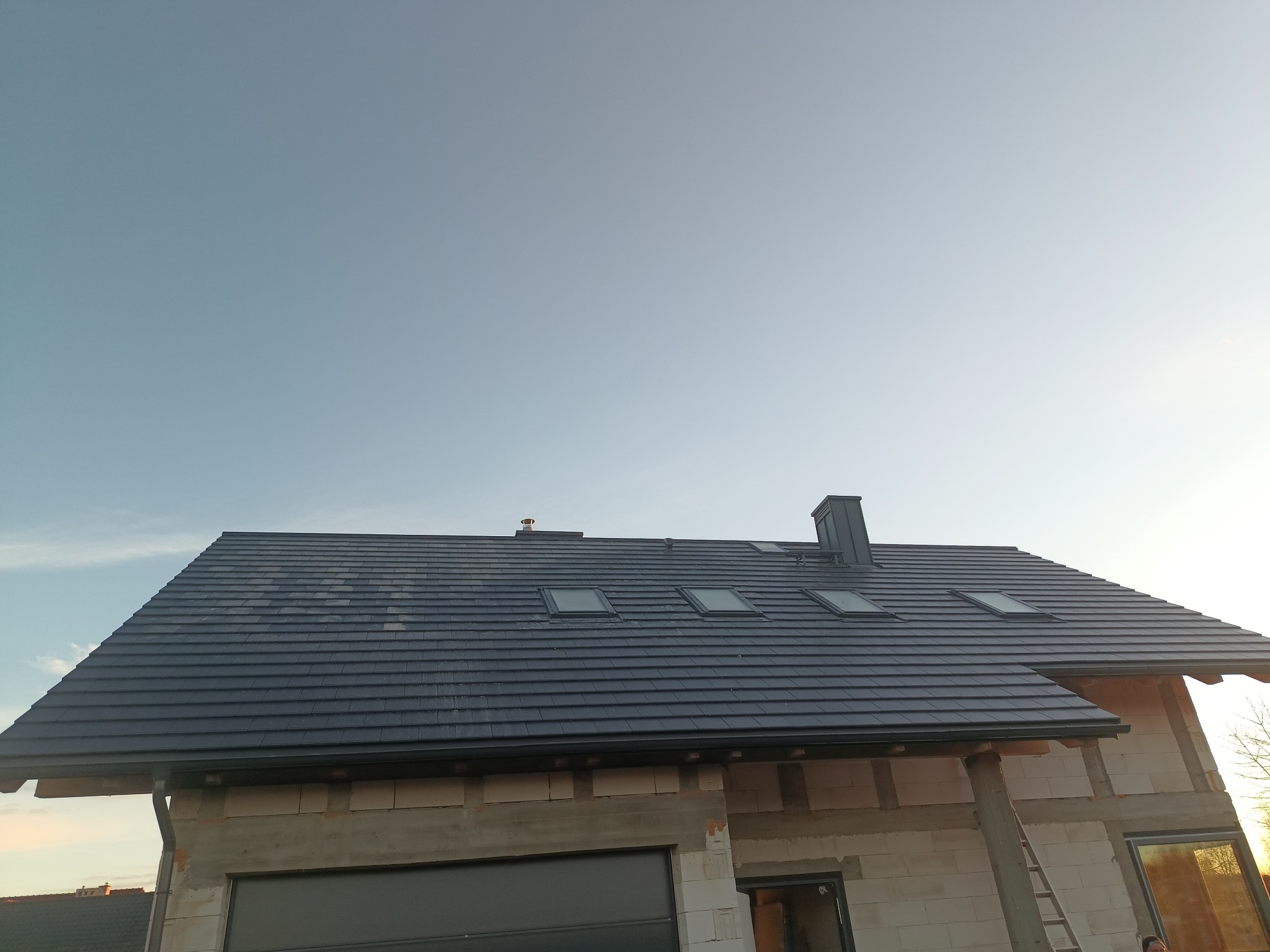 Nowoczesny dom w trakcie budowy, z ciemnoszarą dachówką ceramiczną, oknami dachowymi i kominem, widoczny z dołu, w tle błękitne niebo.