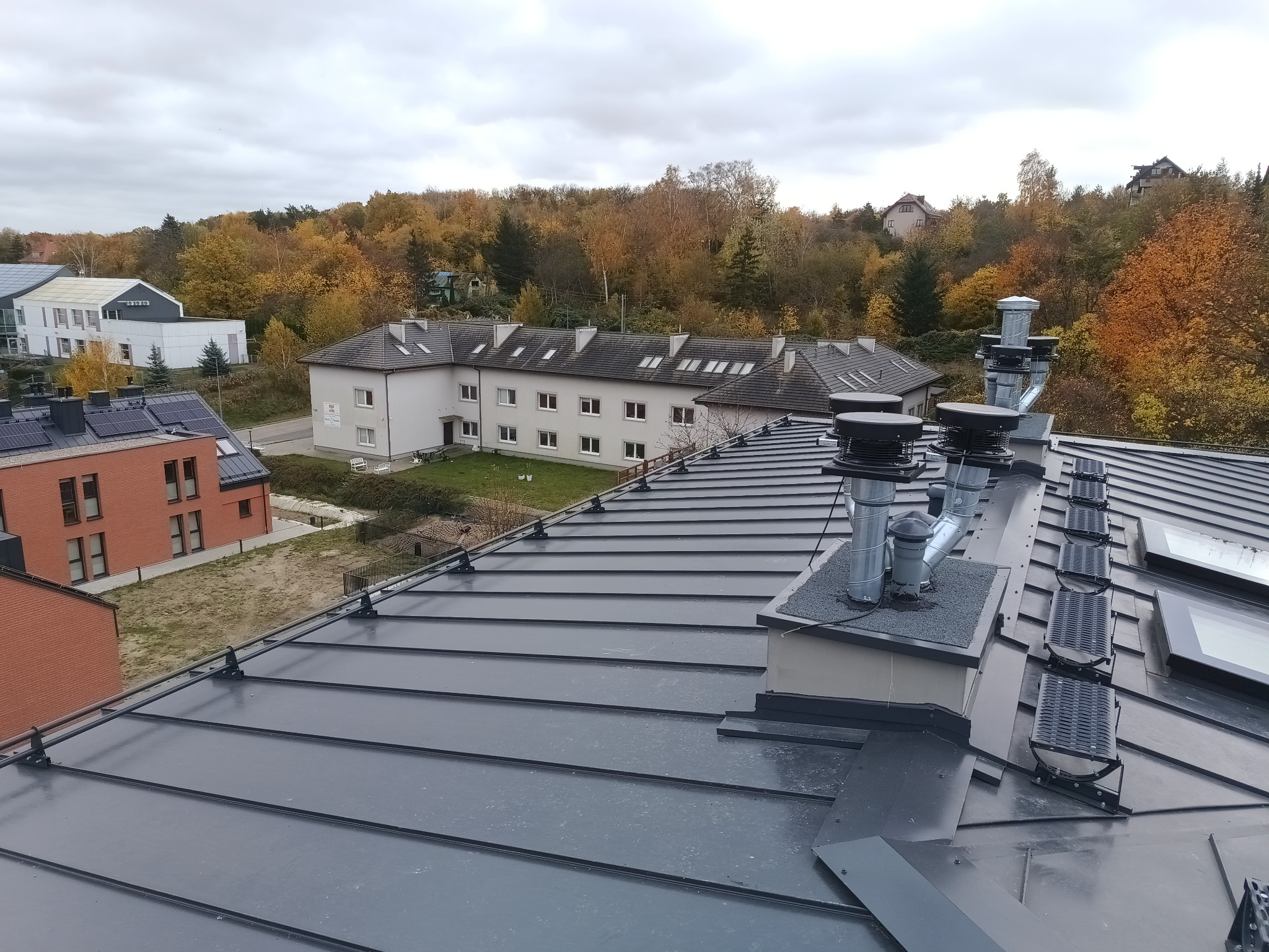 Nowy, ciemnoszary dach z blachy na budynku mieszkalnym, widoczne kominy wentylacyjne i świetliki dachowe, jesienne drzewa w tle.