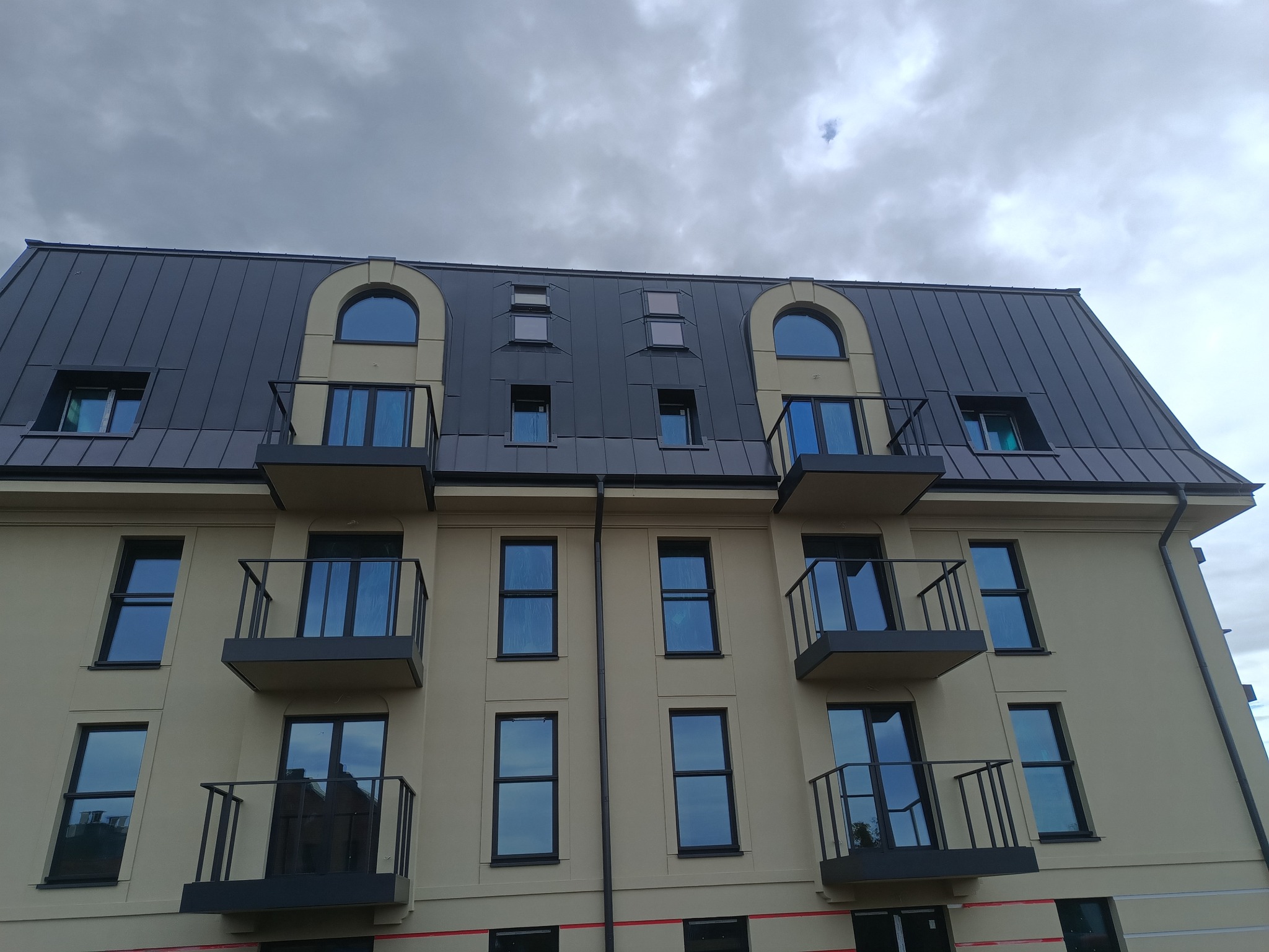 Nowoczesny budynek wielorodzinny z ciemnoszarym dachem z blachy, balkonami i oknami z łukami na poddaszu, uchwycony pod pochmurnym niebem.