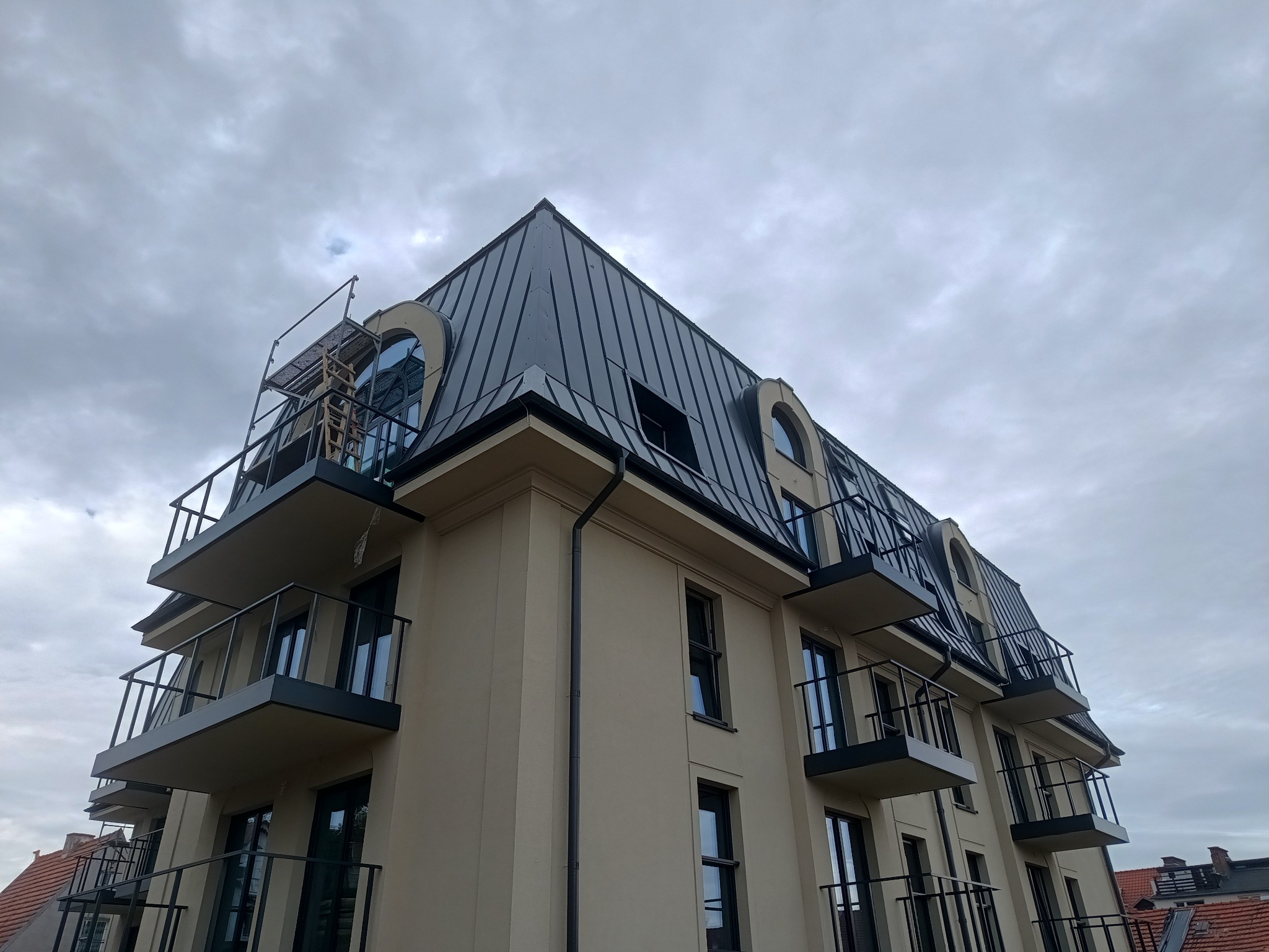 Nowoczesny budynek wielorodzinny z ciemnym, metalowym dachem mansardowym, widoczne balkony z czarnymi balustradami, obróbki blacharskie i rynny w kolorze dachu, na dachu rusztowanie przy oknie.