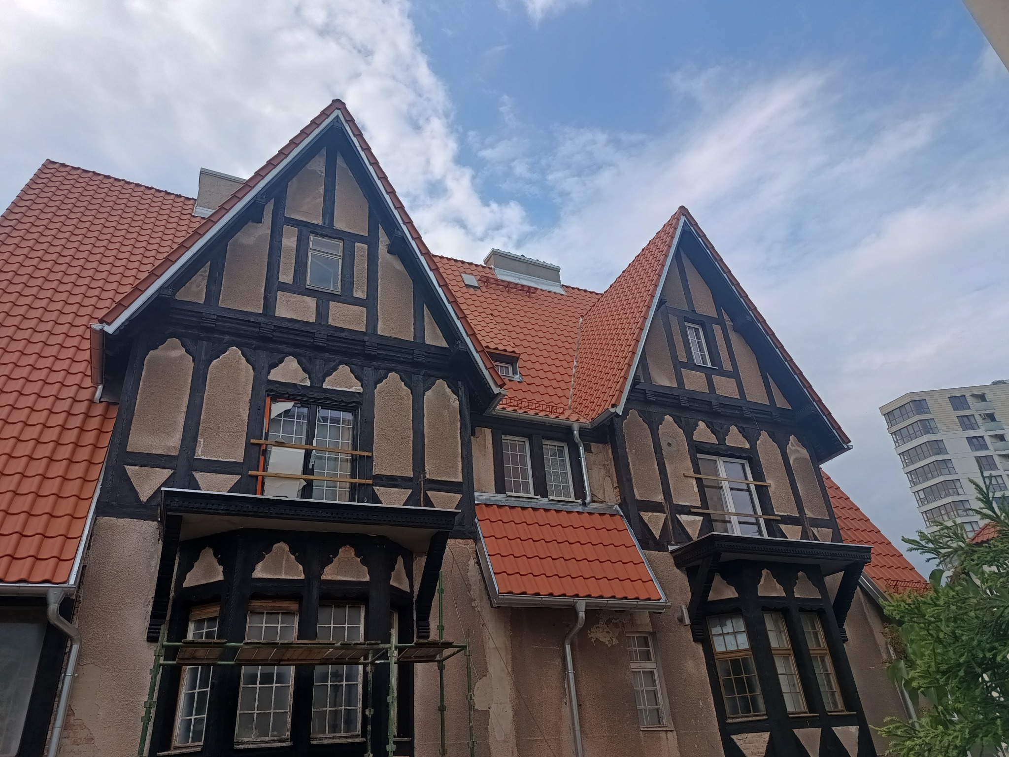 Odnowiony dach z czerwonej dachówki karpiówki na budynku w stylu szachulcowym, widoczne rusztowania i częściowo uszkodzone okna, na tle błękitnego nieba z chmurami.