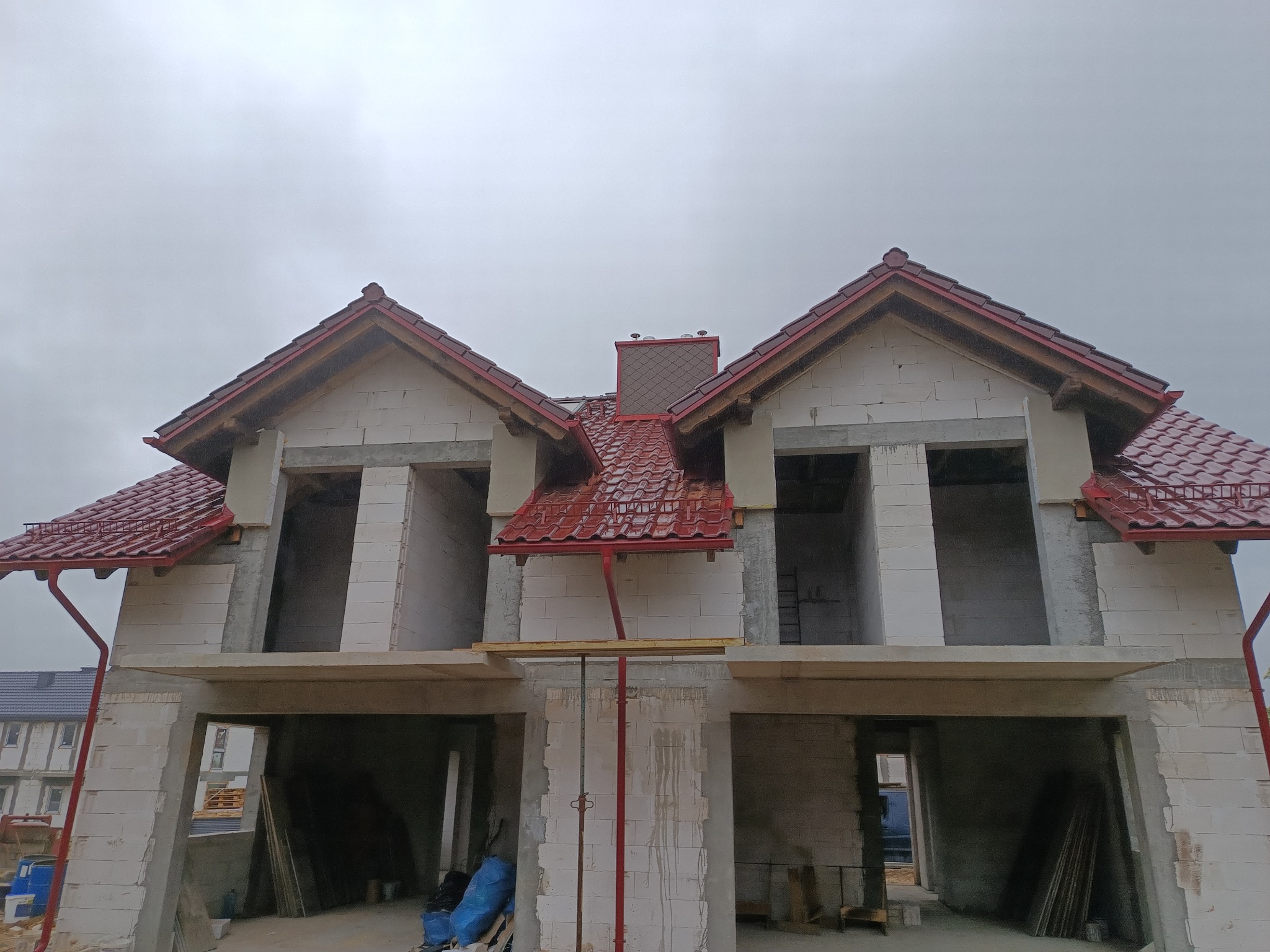 Surowy stan bliźniaka z czerwonym dachem z dachówki ceramicznej i czerwonymi rynnami, widoczne wsporniki podtrzymujące konstrukcję.