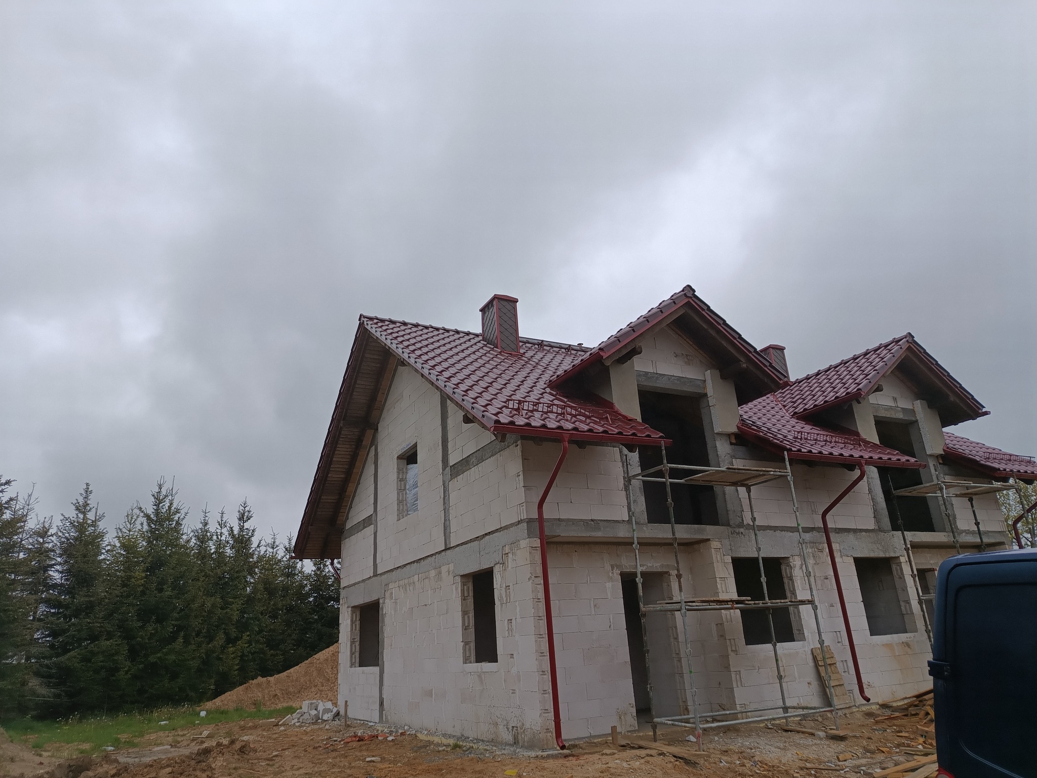 Budowa domu w Sierakowicach: widok na budynek w stanie surowym z częściowo położonym czerwonym dachem i zamontowanymi rynnami w tym samym kolorze, na tle pochmurnego nieba i drzew.