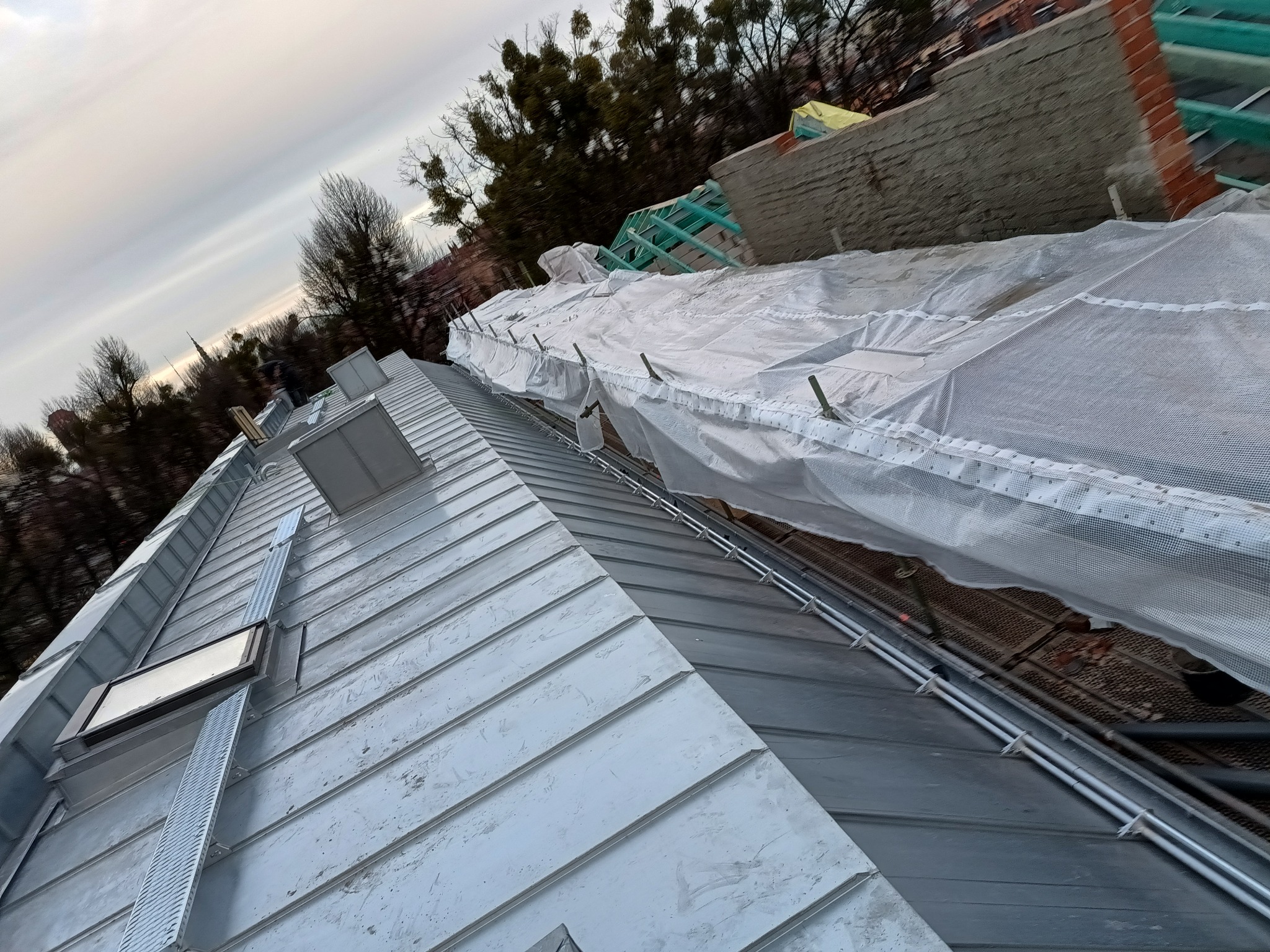 Metalowy dach z oknami dachowymi i zabezpieczeniem z siatki, widoczne elementy konstrukcji dachu i fragment murowanej ściany szczytowej w tle, pochmurne niebo.