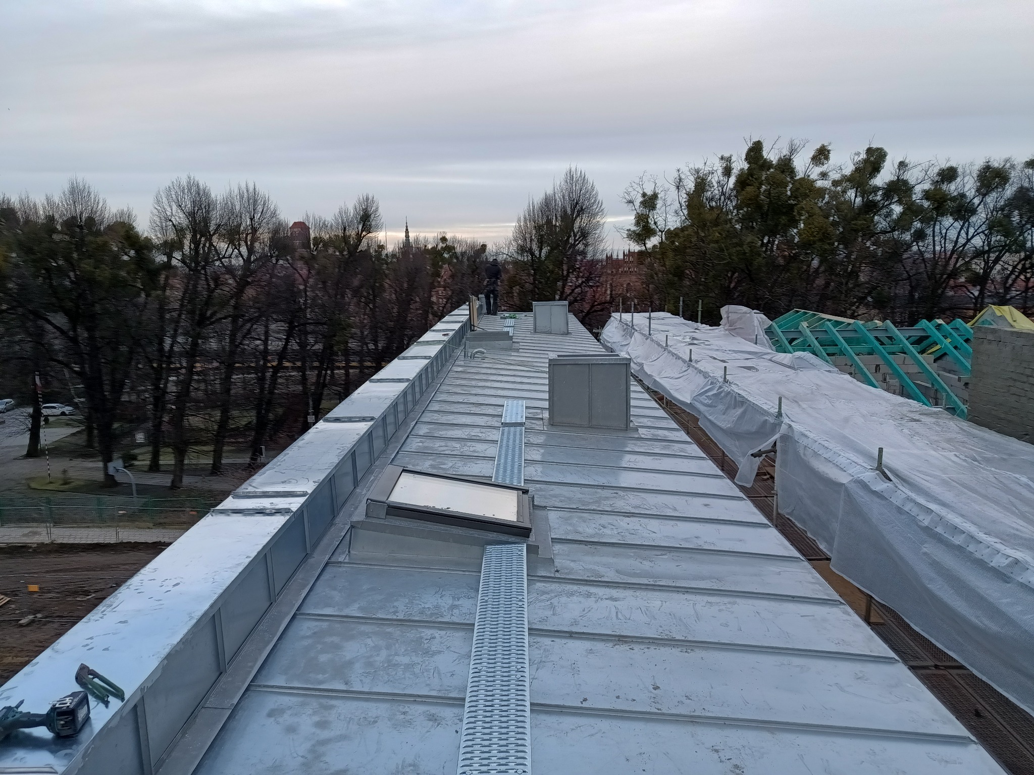 Nowy dach z blachy na budynku miejskim, widoczne obróbki blacharskie, okno dachowe, kominy i zabezpieczenia, w tle drzewa i zabudowa miejska.