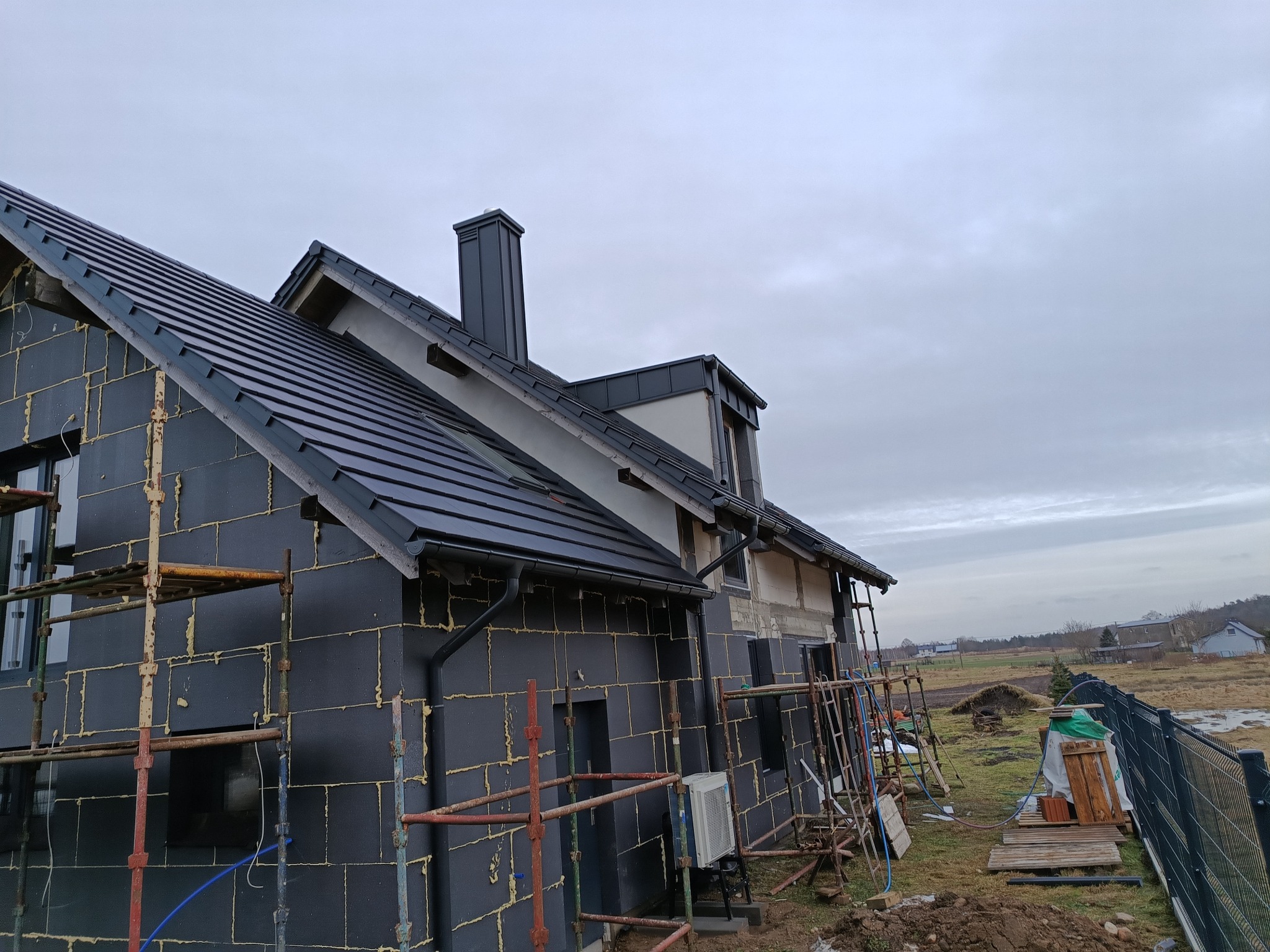 Dom w trakcie budowy, z czarną izolacją ścian i nowym, ciemnym dachem z rynnami, widoczne rusztowania i elementy wykończeniowe.