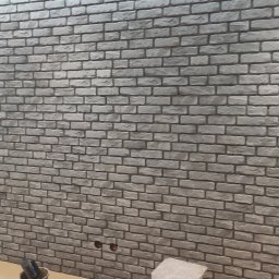 Arkadiusz Kozłowski Usługi Glazurnicze - Ściana wykończona szarą cegłą dekoracyjną w trakcie prac, widoczne gniazdka elektryczne i narzędzia w wiadrze na podłodze.