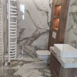 Nowoczesna łazienka z marmurowymi płytkami, podświetlanym sufitem, grzejnikiem drabinkowym i drewnianym elementem zabudowy z półkami.