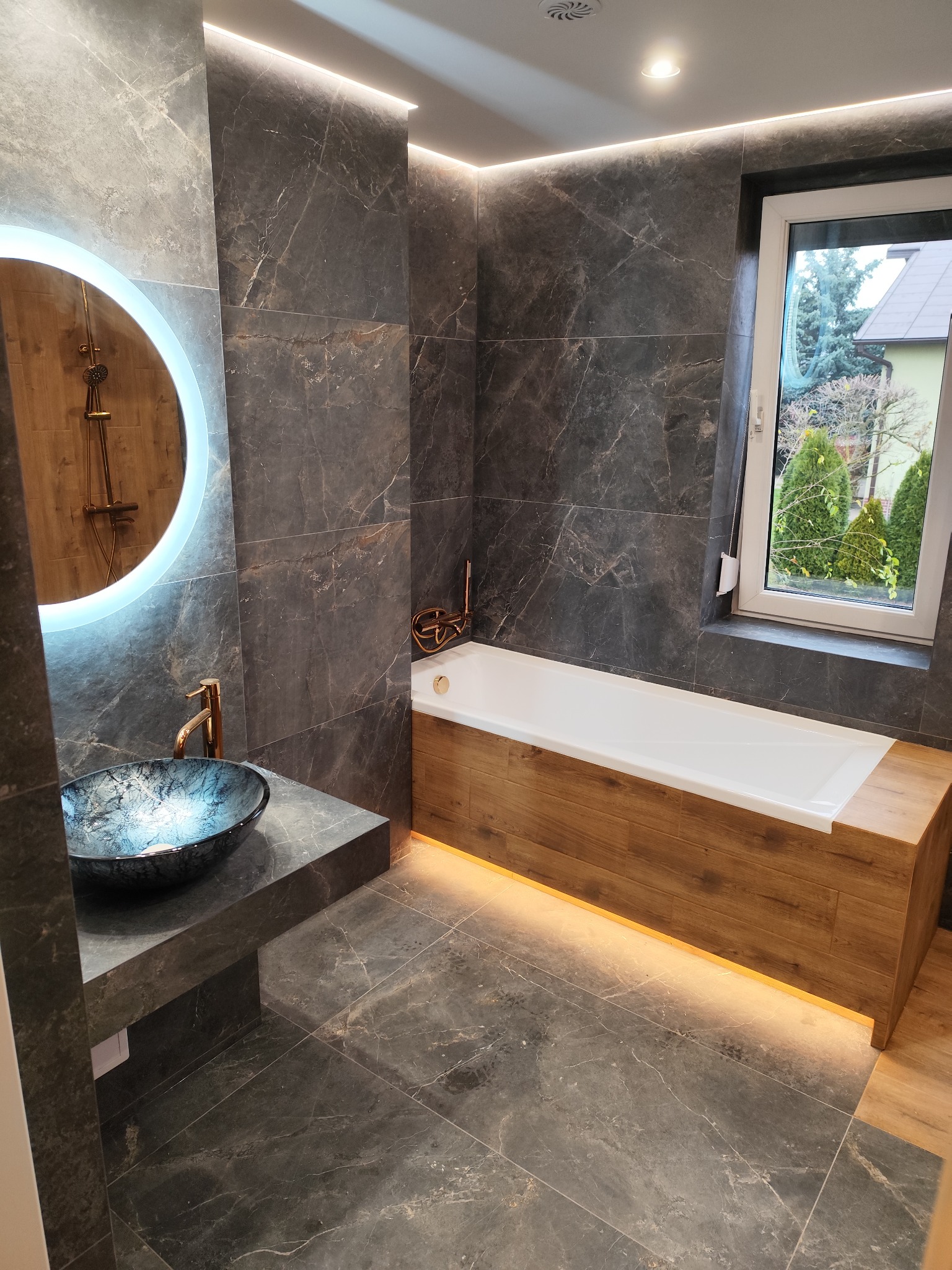 Nowoczesna łazienka z szarymi płytkami imitującymi marmur, okrągłym lustrem z podświetleniem LED, umywalką w kolorze granatowym z miedzianą armaturą oraz wanną obudowaną drewnem z podświetleniem...