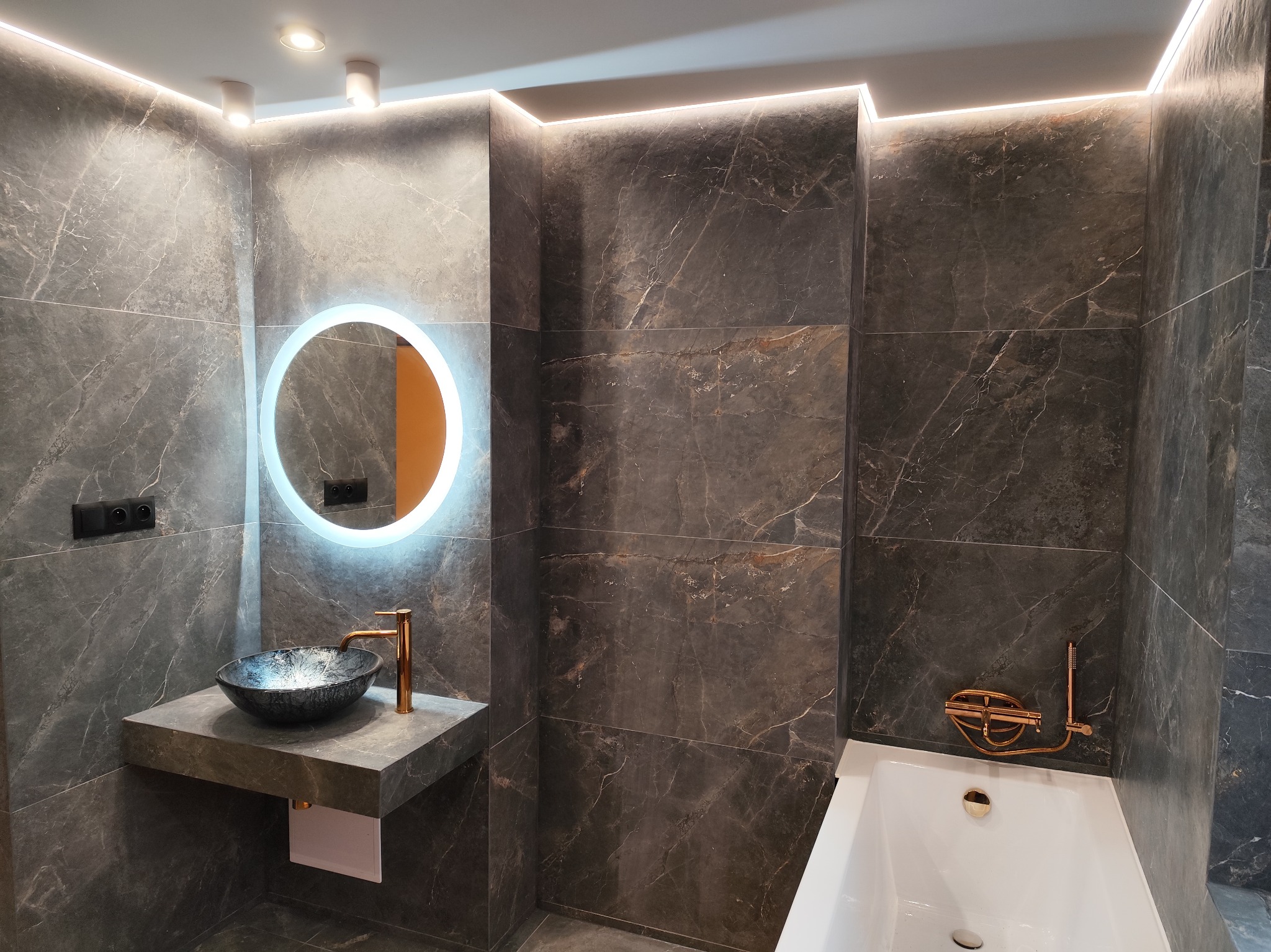 Nowoczesna łazienka z szarymi marmurowymi płytkami, okrągłym podświetlanym lustrem, nablatową umywalką w kolorze ciemnego granitu z złotą baterią i białą wanną z złotą armaturą.