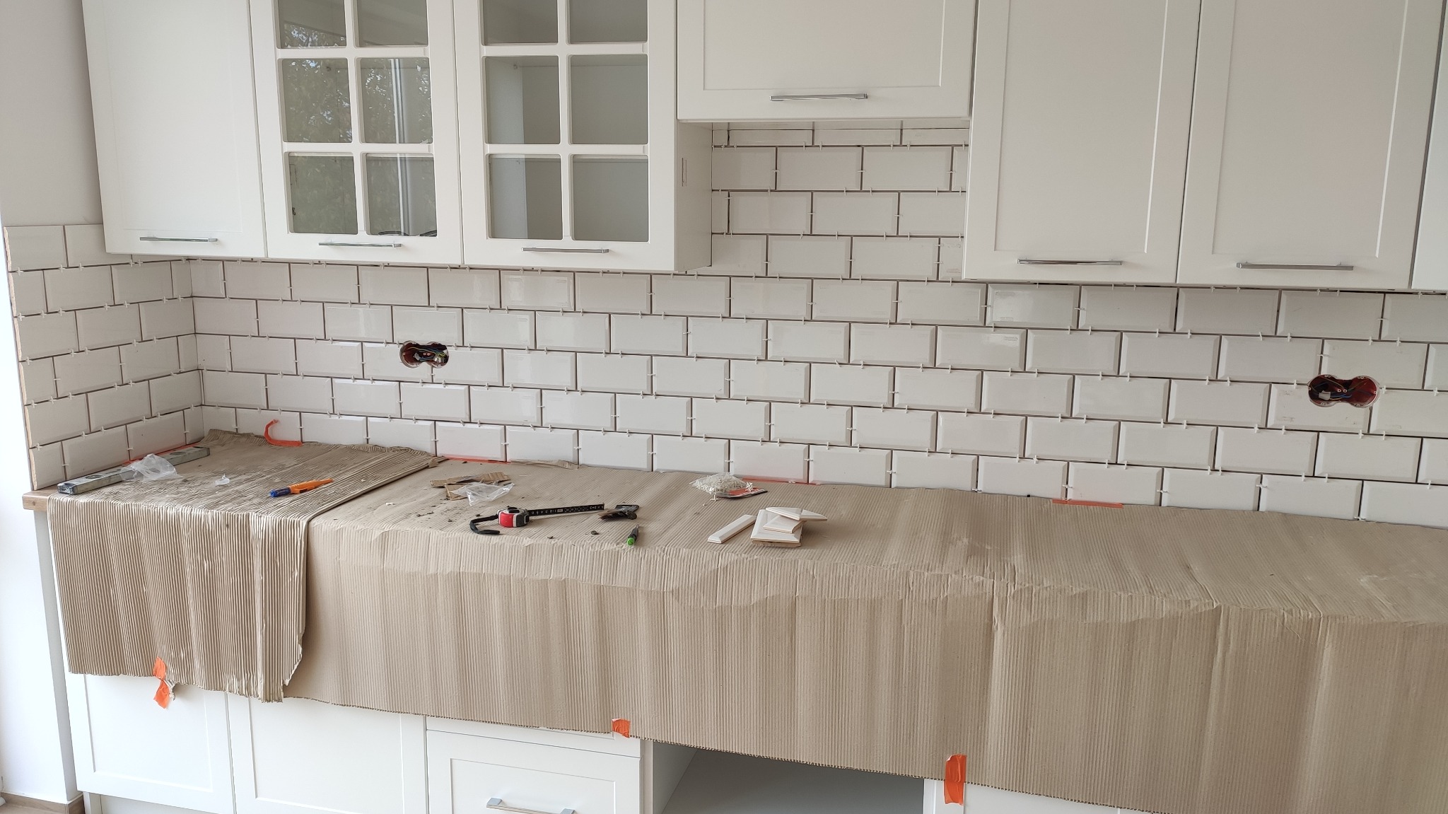 Białe, prostokątne płytki ułożone na ścianie w kuchni, zabezpieczony blat roboczy tekturą falistą, widoczne narzędzia i gniazdko elektryczne bez osprzętu.