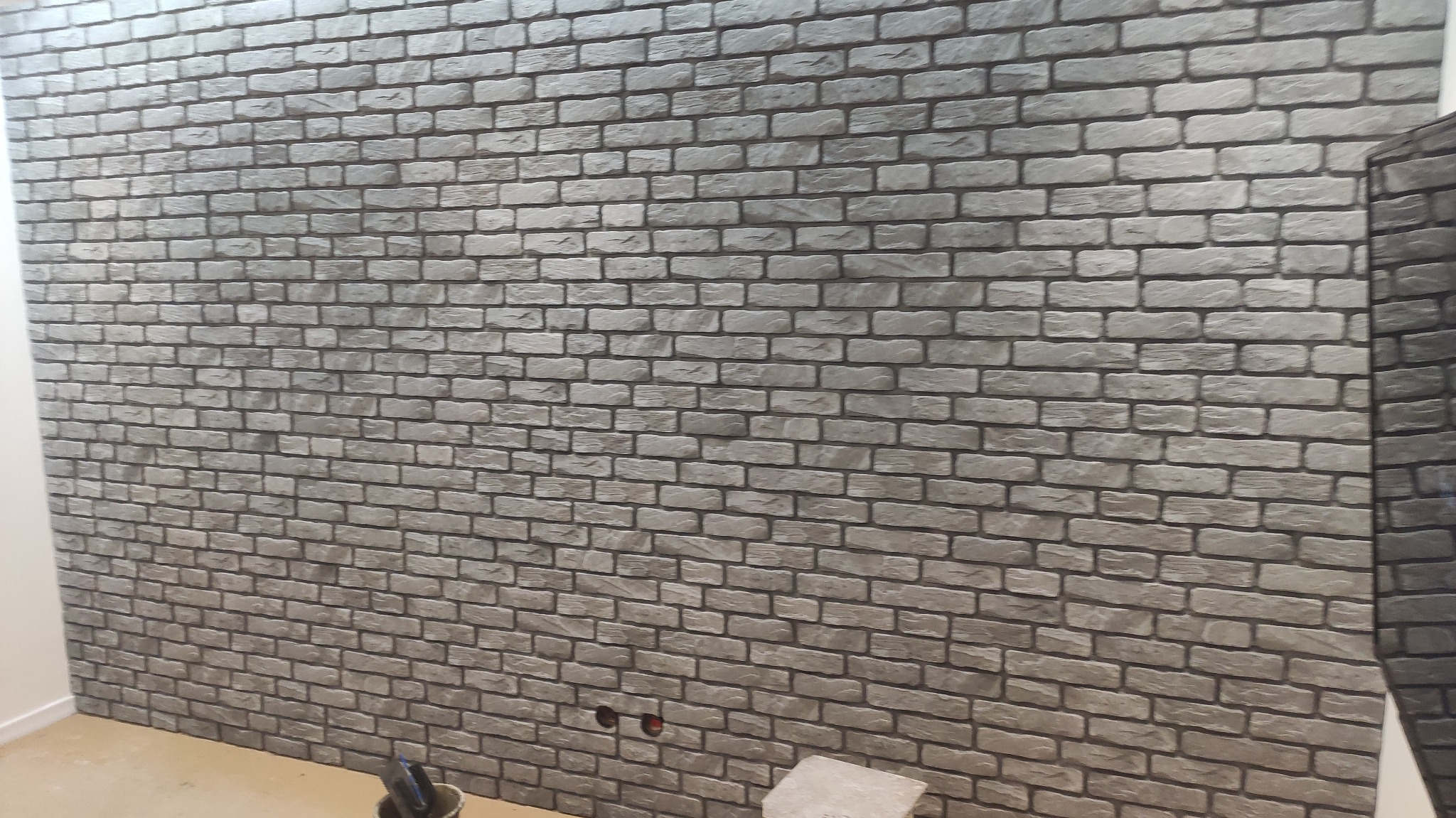 Ściana wykończona szarą cegłą dekoracyjną w trakcie prac, widoczne gniazdka elektryczne i narzędzia w wiadrze na podłodze.