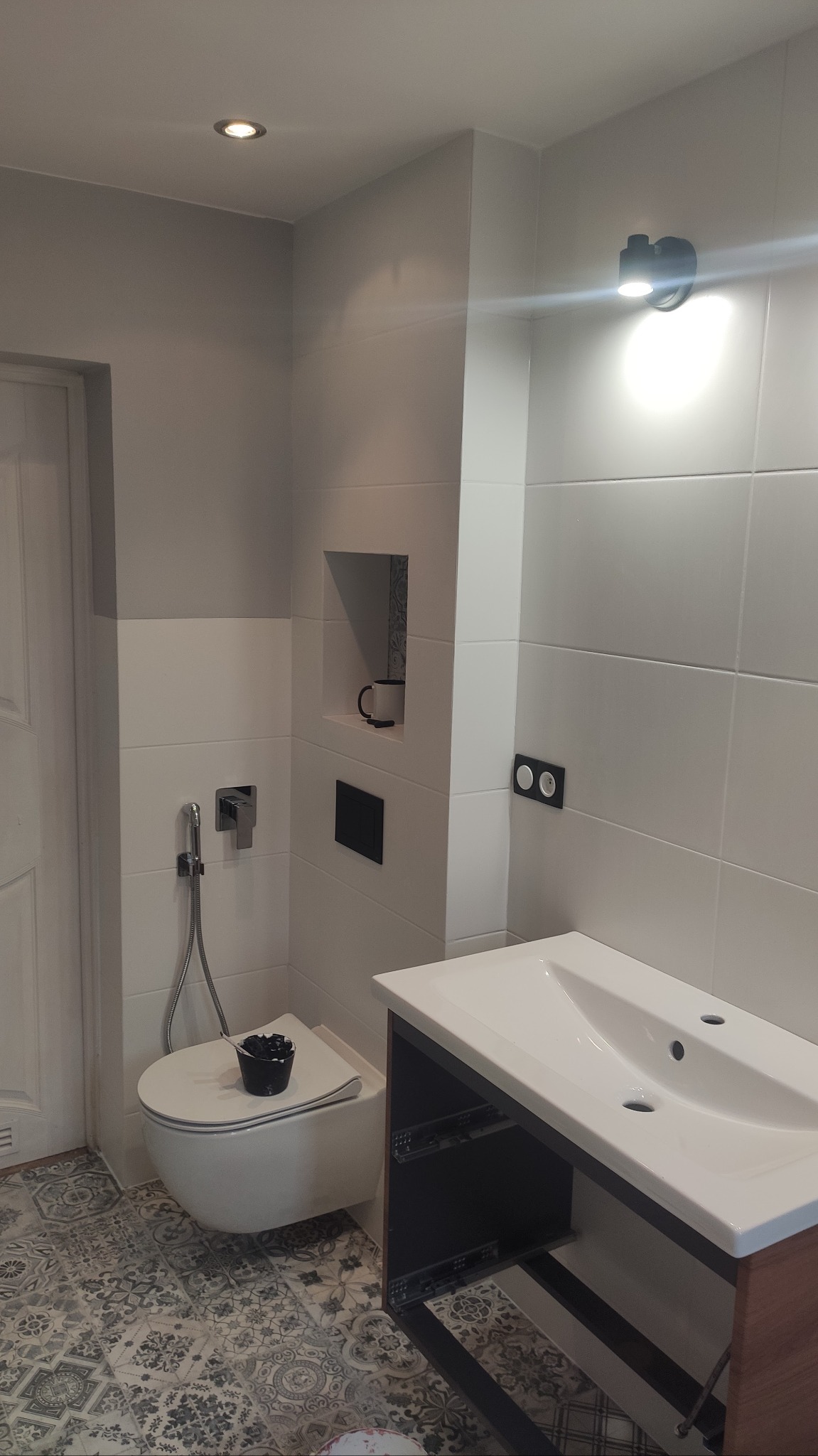 Nowoczesna łazienka po remoncie z białą armaturą, płytkami w odcieniach szarości i wzorzystą podłogą. Na spłuczce toalety stoi czarny pojemnik.